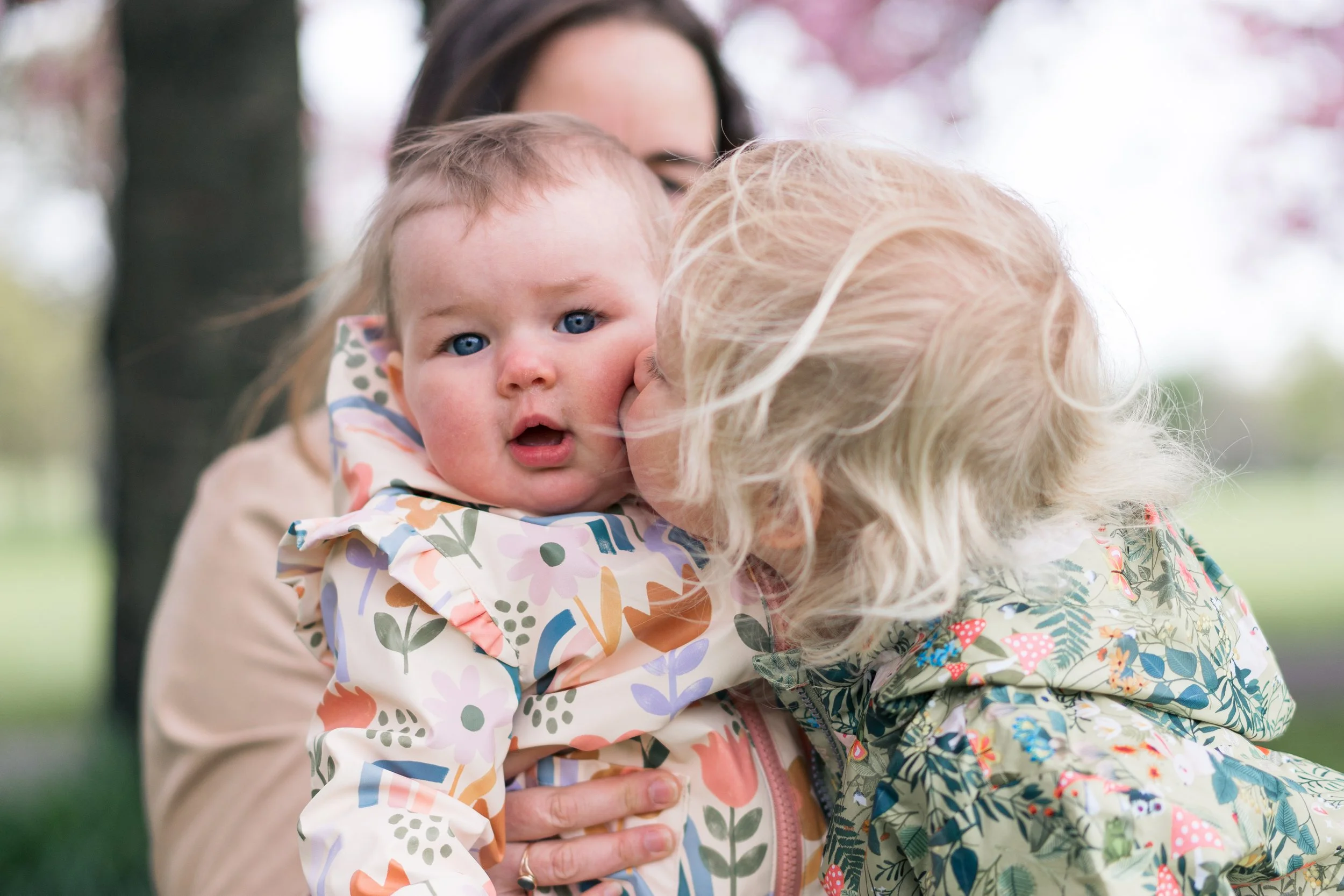 A young child with blue eyes and a surprised expression, being kissed on the cheek by an older woman with blond hair, outdoors with pink blossoms and green background.