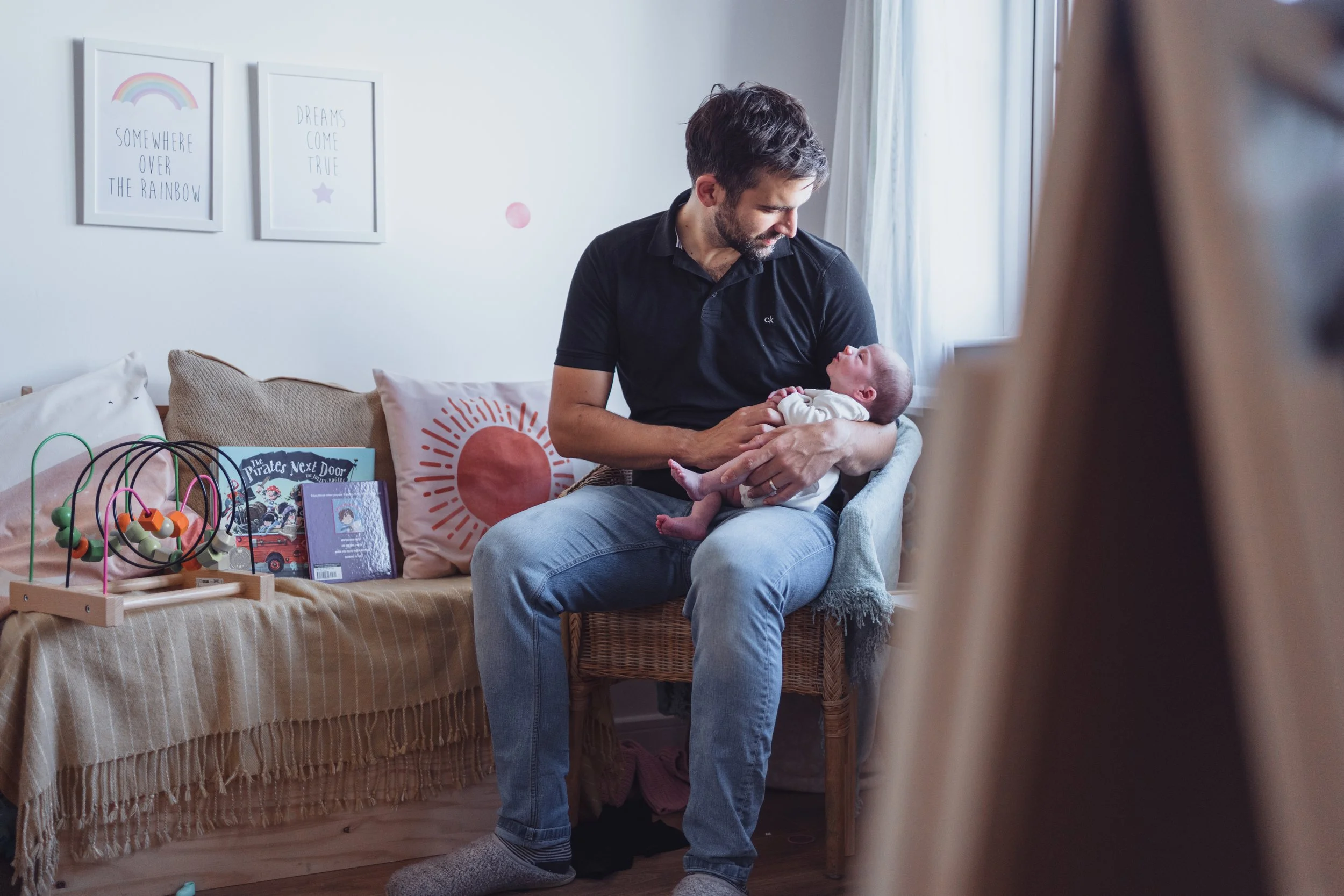 A man holding a baby inside a home, sitting on a wicker chair near a couch with books and toys, and decorative wall art.