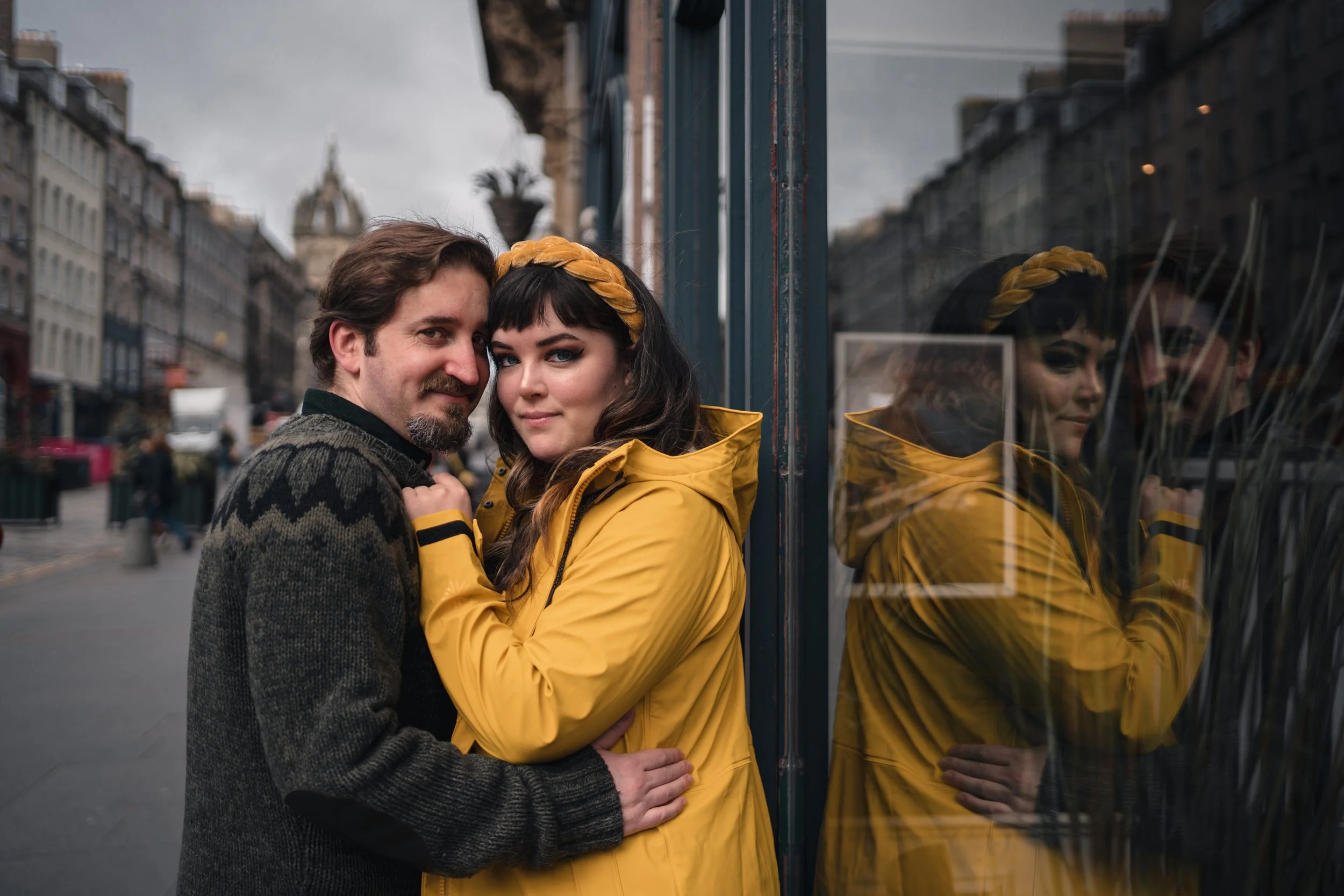 A young woman in a yellow raincoat and headband smiling with a man in a dark sweater, both standing against a reflective glass wall on a city street.