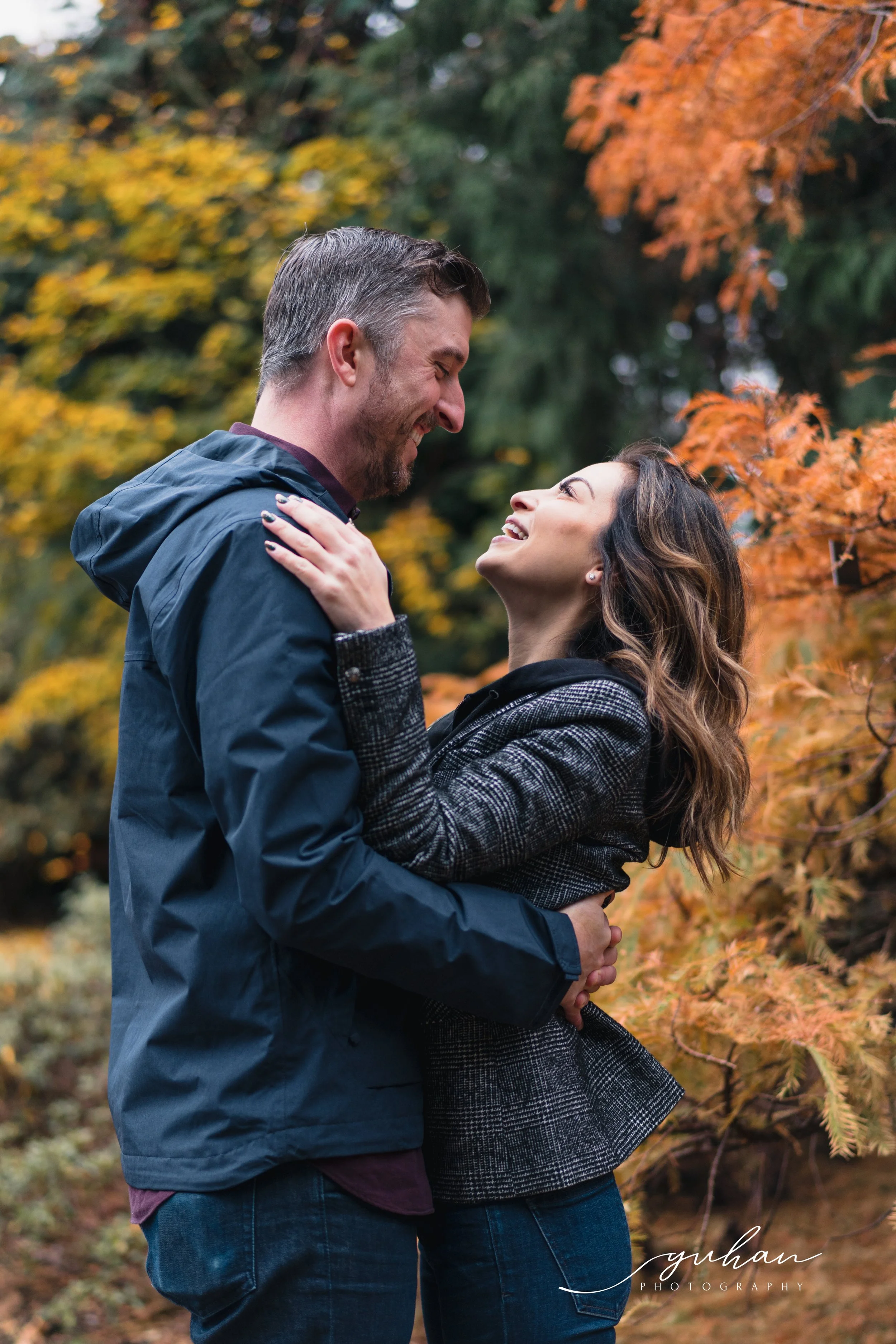 A couple embraces outdoors during autumn, surrounded by orange and yellow fall foliage, smiling at each other.