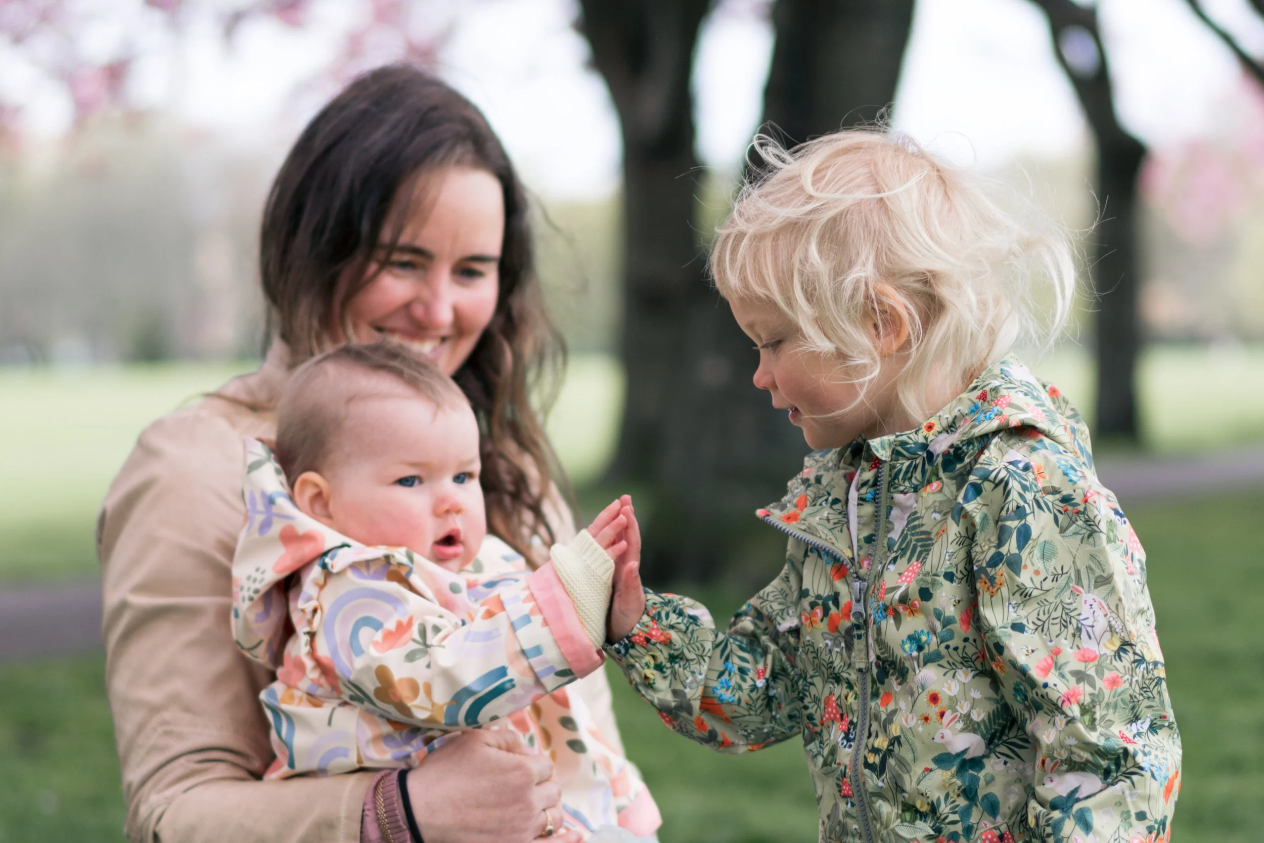 A woman holds a baby while a young girl gives them a high-five outdoors in a park with green grass and trees.