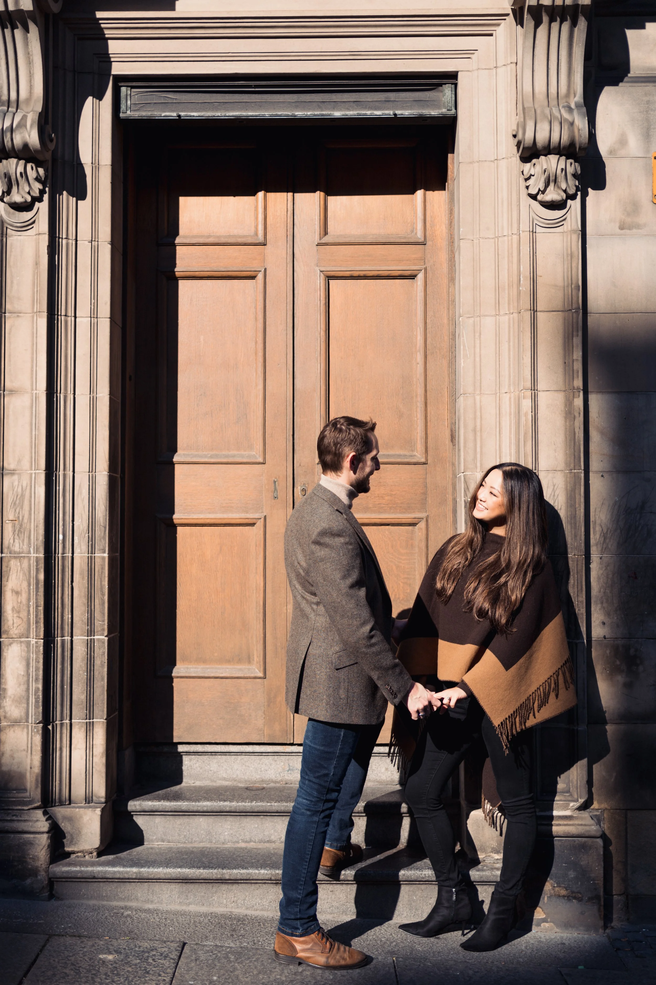 A man and woman holding hands, standing and smiling at each other on steps outside a building with large wooden doors.