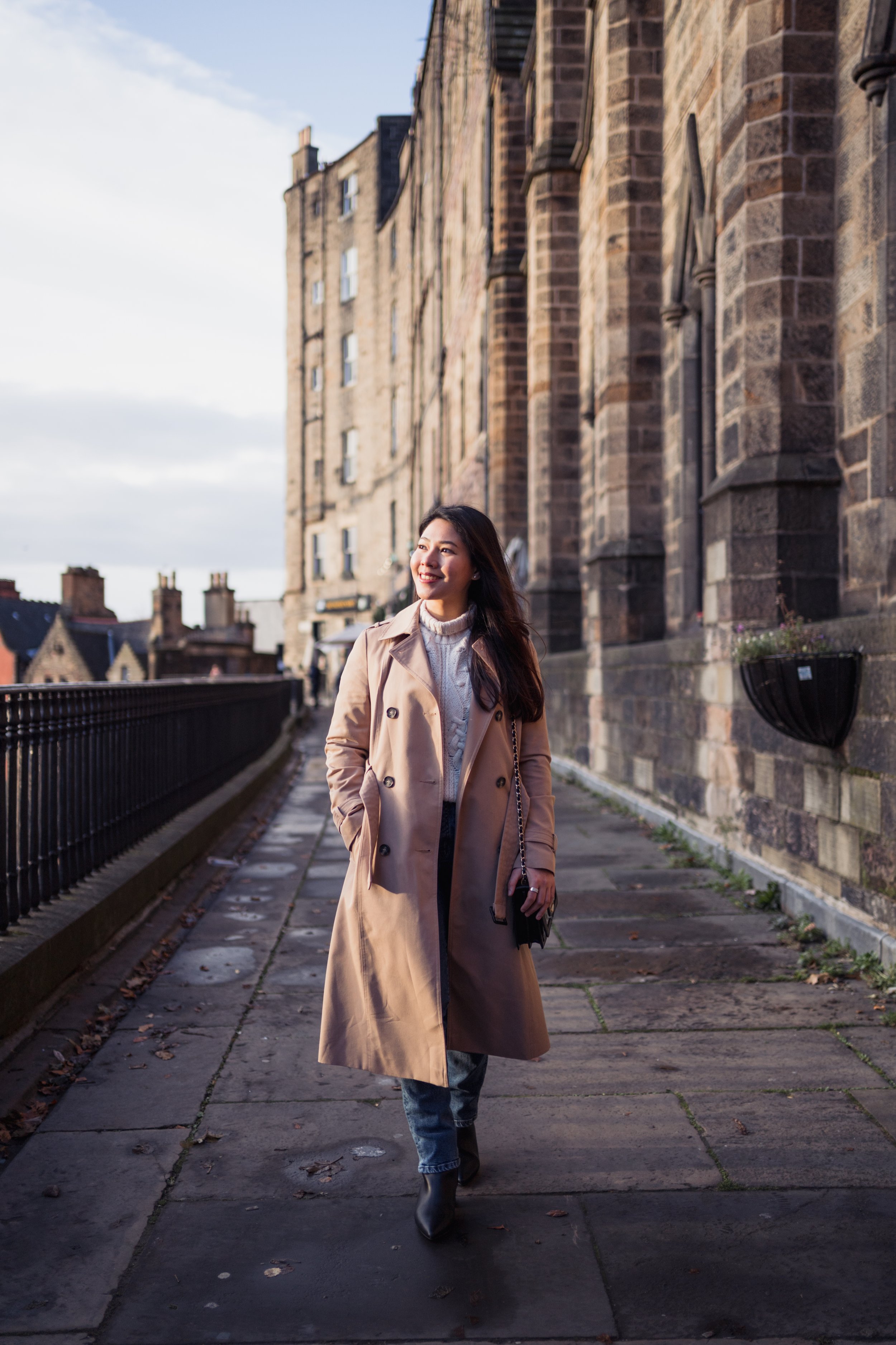 A woman walking on a sidewalk in front of an old stone building with potted plants on the wall, smiling as sunlight illuminates her face.
