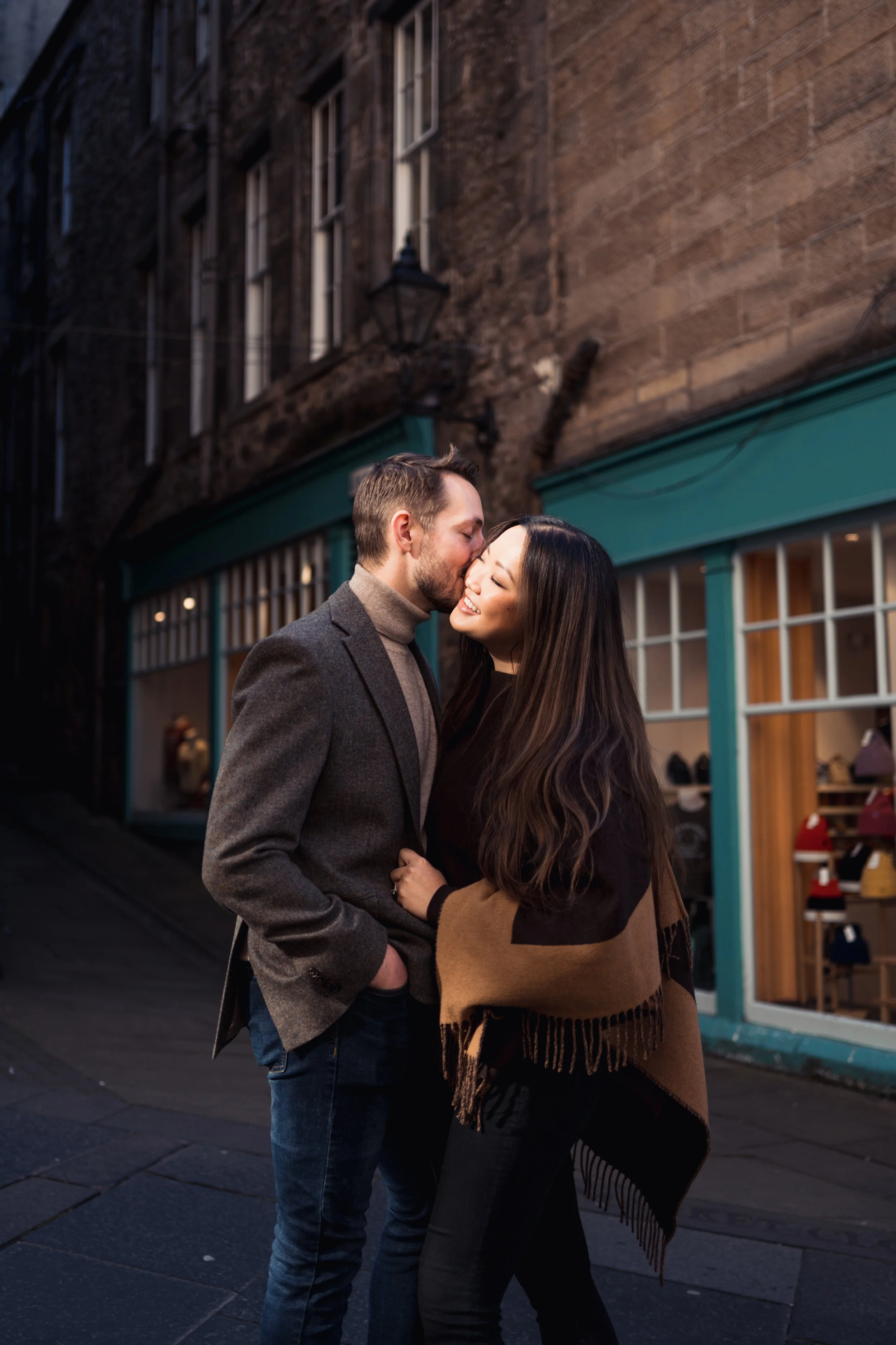 A young man and woman share a kiss on a city street at dusk. She is smiling with eyes closed, wrapped in a brown and black shawl, and he is wearing a brown blazer over a gray turtleneck. Behind them are storefronts with teal trim and a stone building