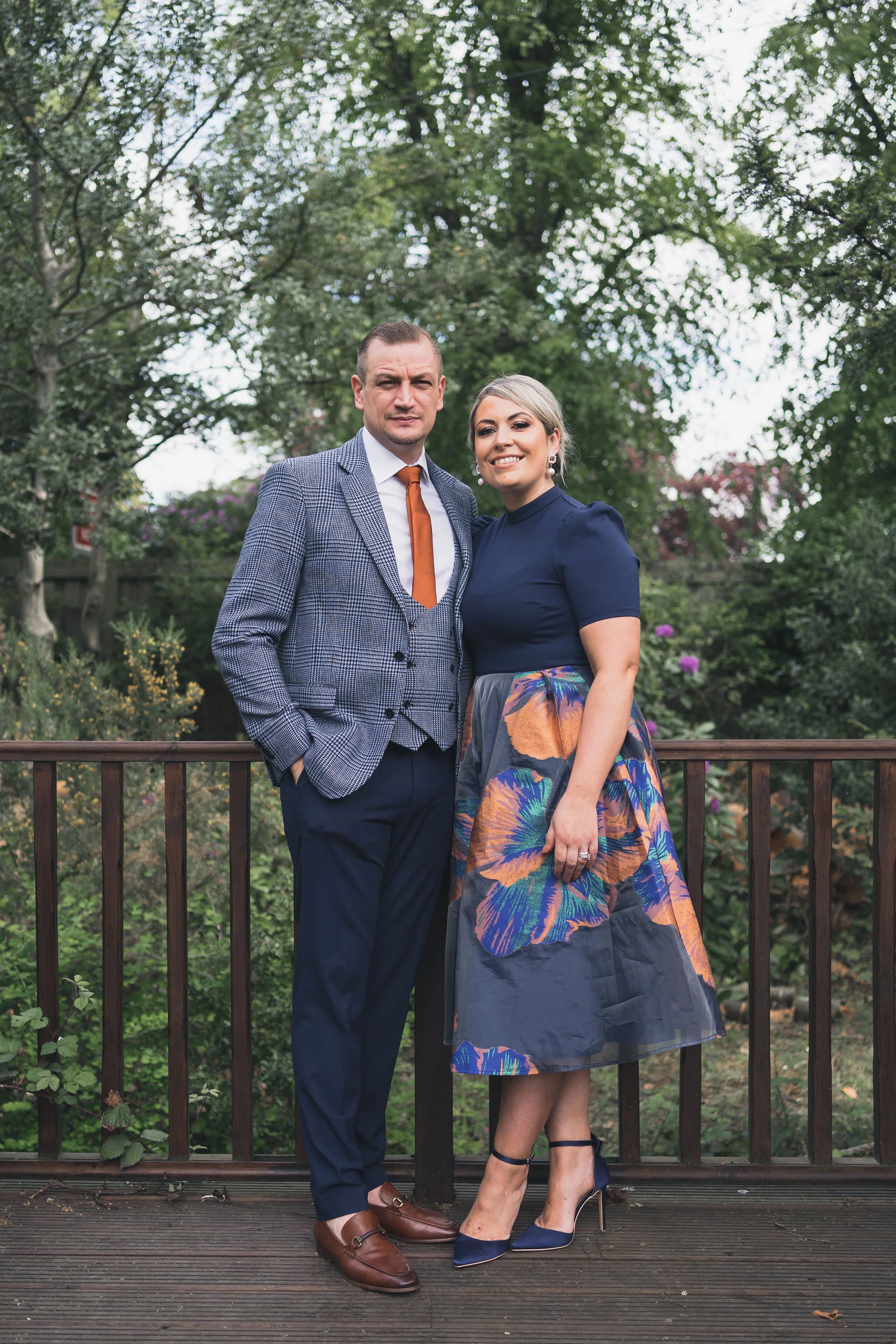 A man and woman stand close together outdoors on a wooden deck, surrounded by green trees and bushes, dressed in semi-formal attire, smiling at the camera.
