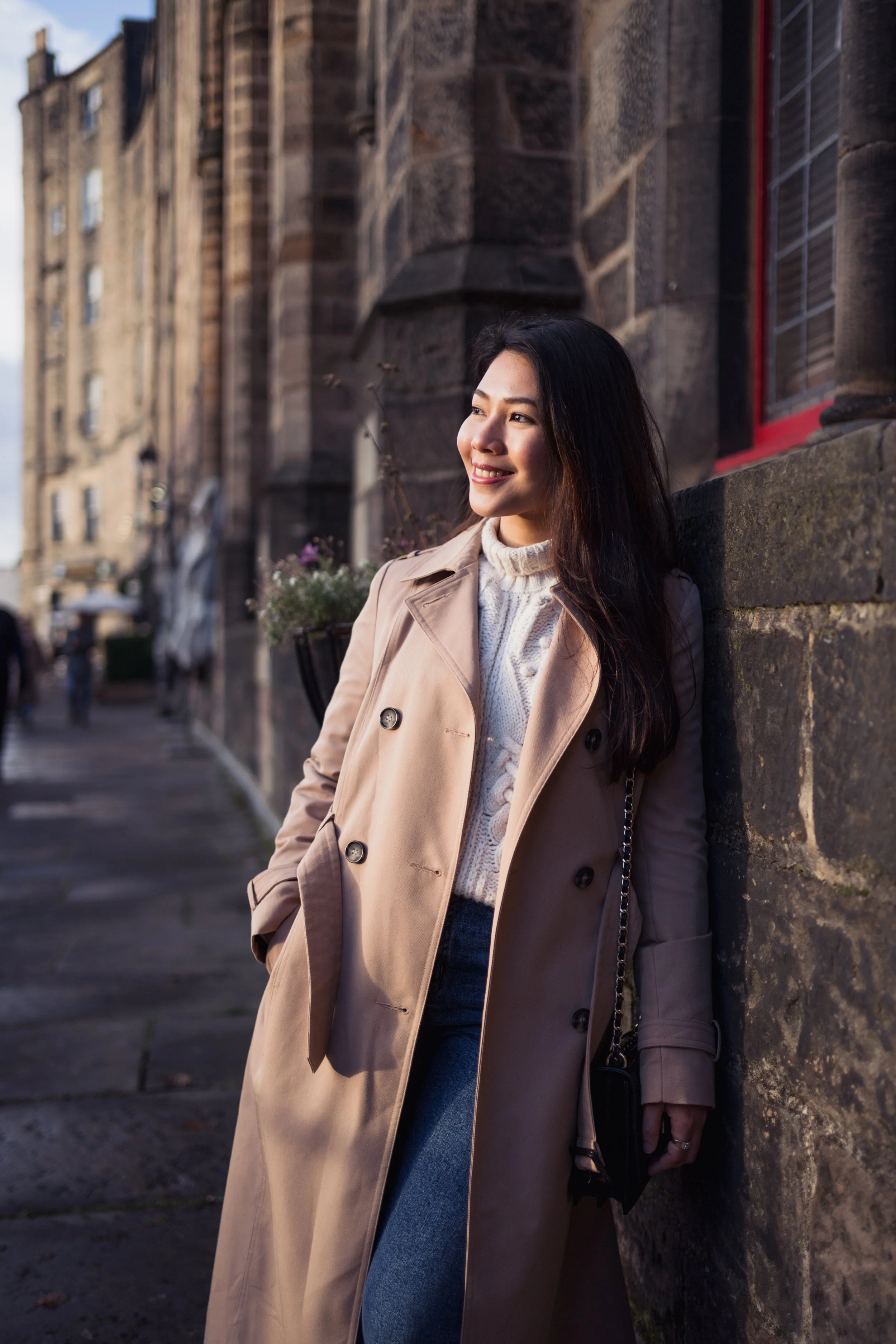 A young woman with long dark hair smiling and leaning against a stone wall in a city with historic buildings, wearing a beige trench coat and a white sweater.