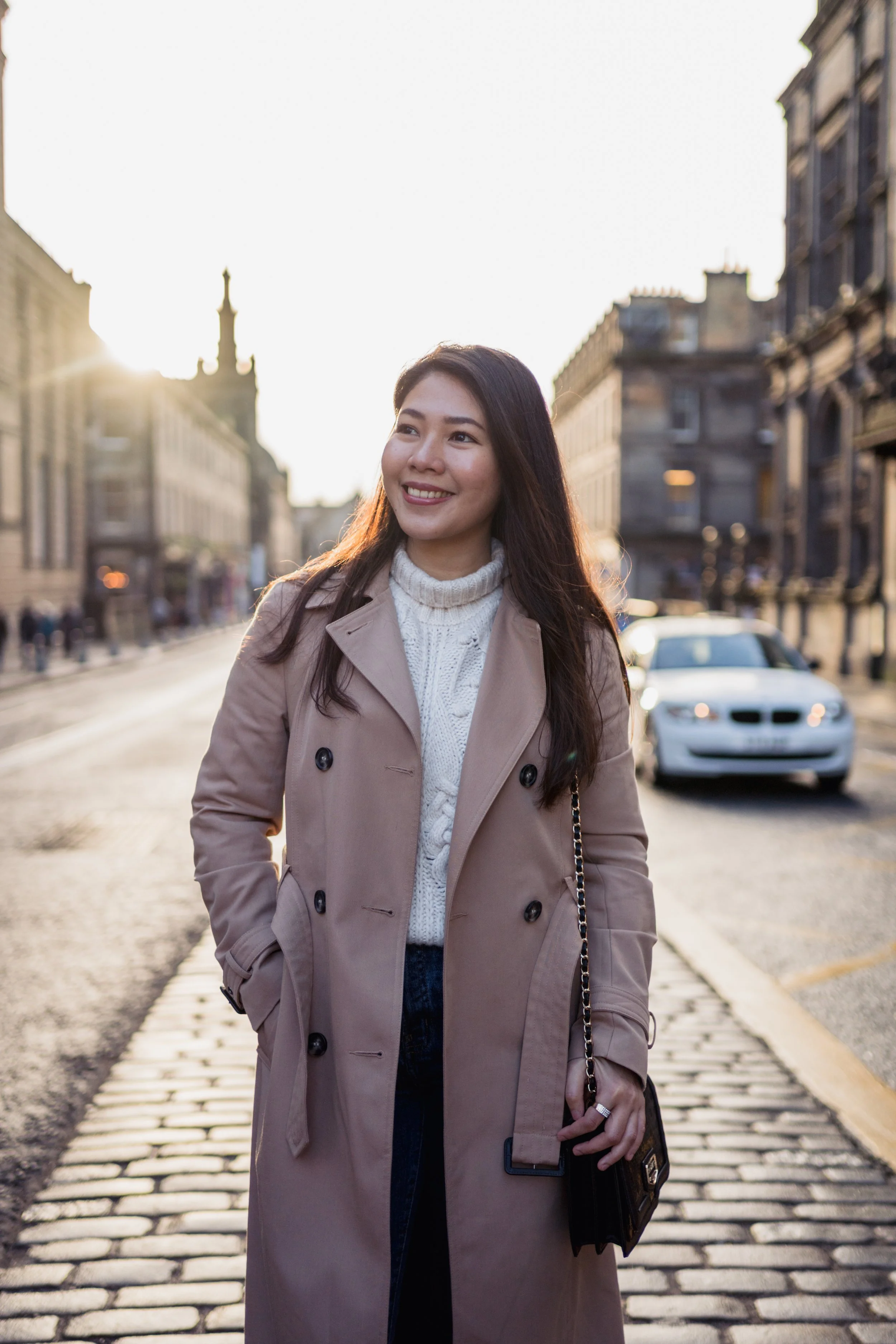 A young woman in a beige trench coat and white sweater smiling on a city street during sunset, with cars and historic buildings in the background.
