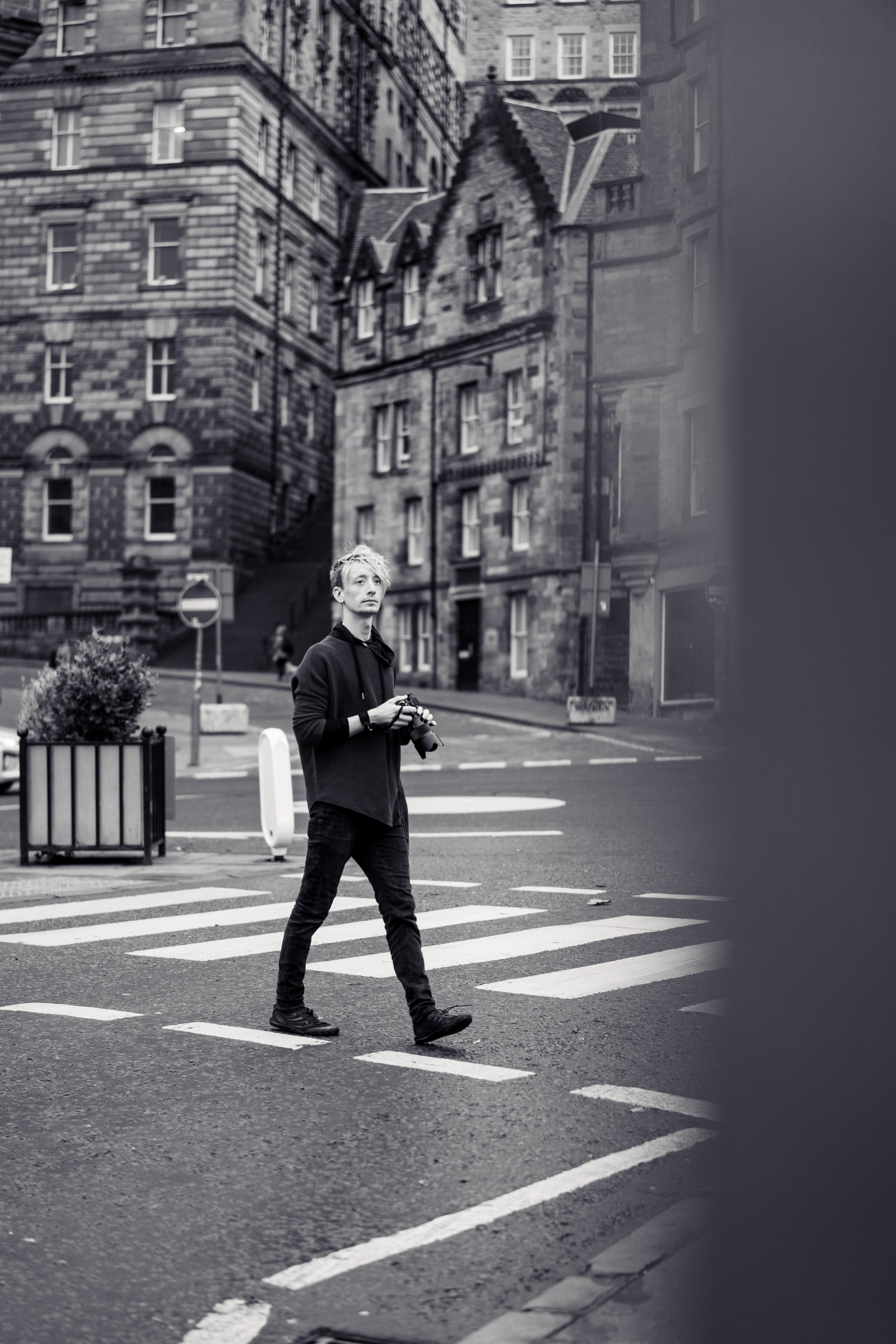 A young man walks across a crosswalk in an urban area, holding a camera and looking to the side, with tall historic stone buildings in the background. The scene is in black and white.