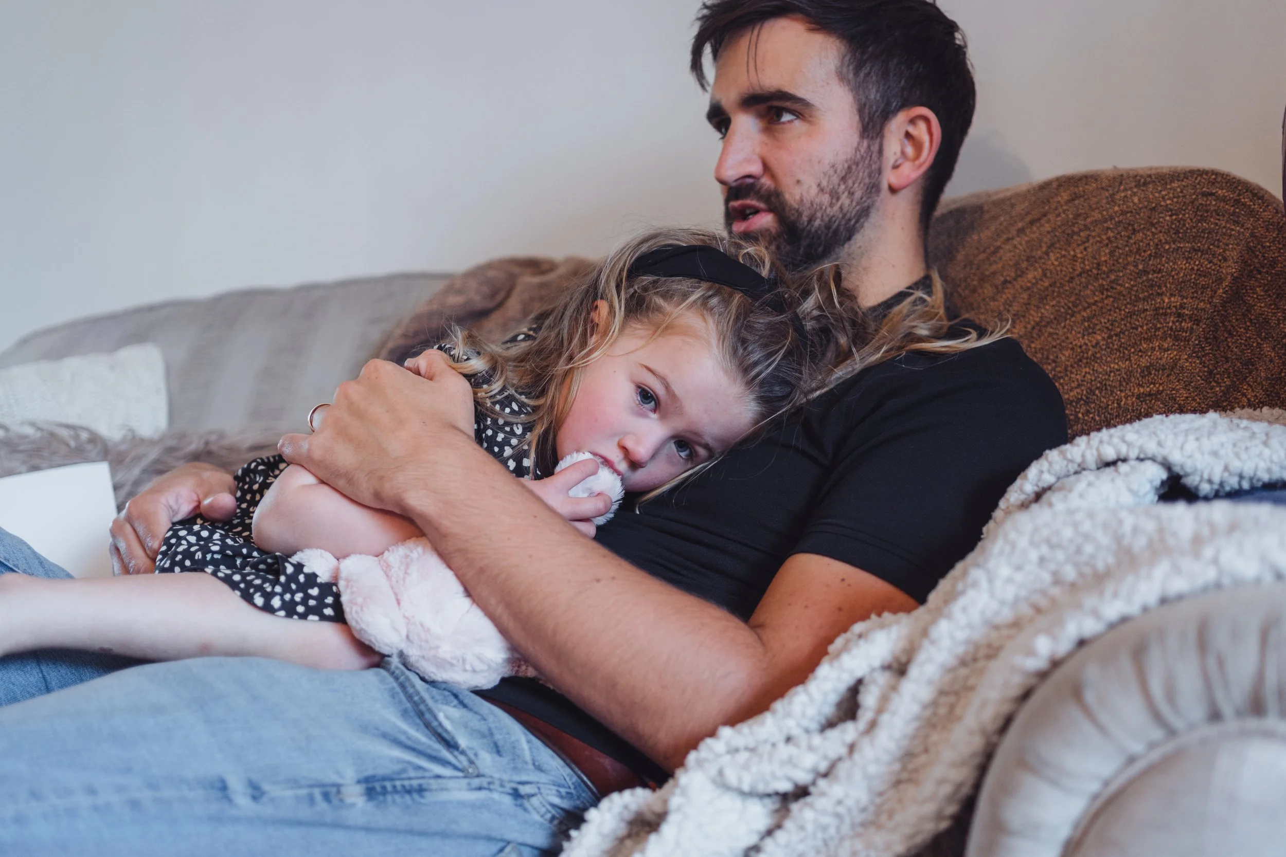 A man with a beard and a young girl with curly blonde hair and a black headband lying on a couch. The girl is resting her head on the man's chest, covering part of his shirt and looking at the camera. The man is looking away, with his arm around the 
