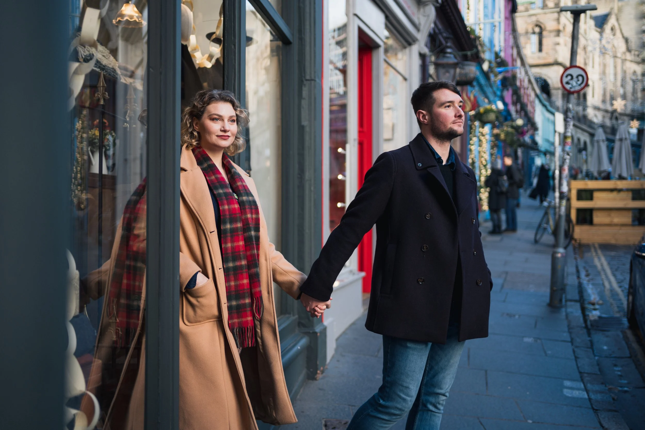 A man and woman holding hands on a city sidewalk in front of shop windows, with the woman standing inside and the man outside, both wearing warm coats.