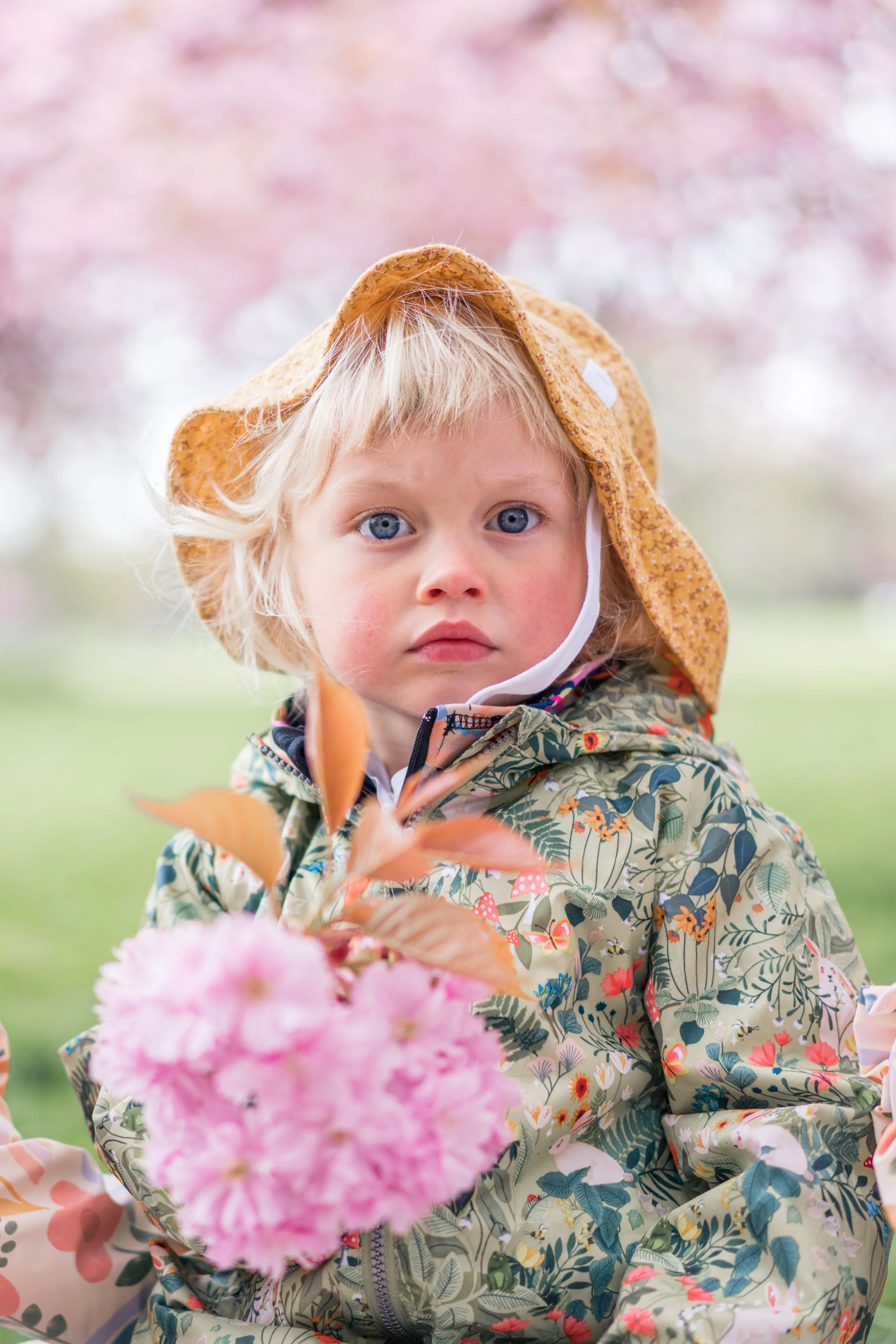 A young child with blonde hair and blue eyes, wearing a yellow hat and a patterned jacket, holds pink flowers outdoors, with pink blossoms in the blurred background.