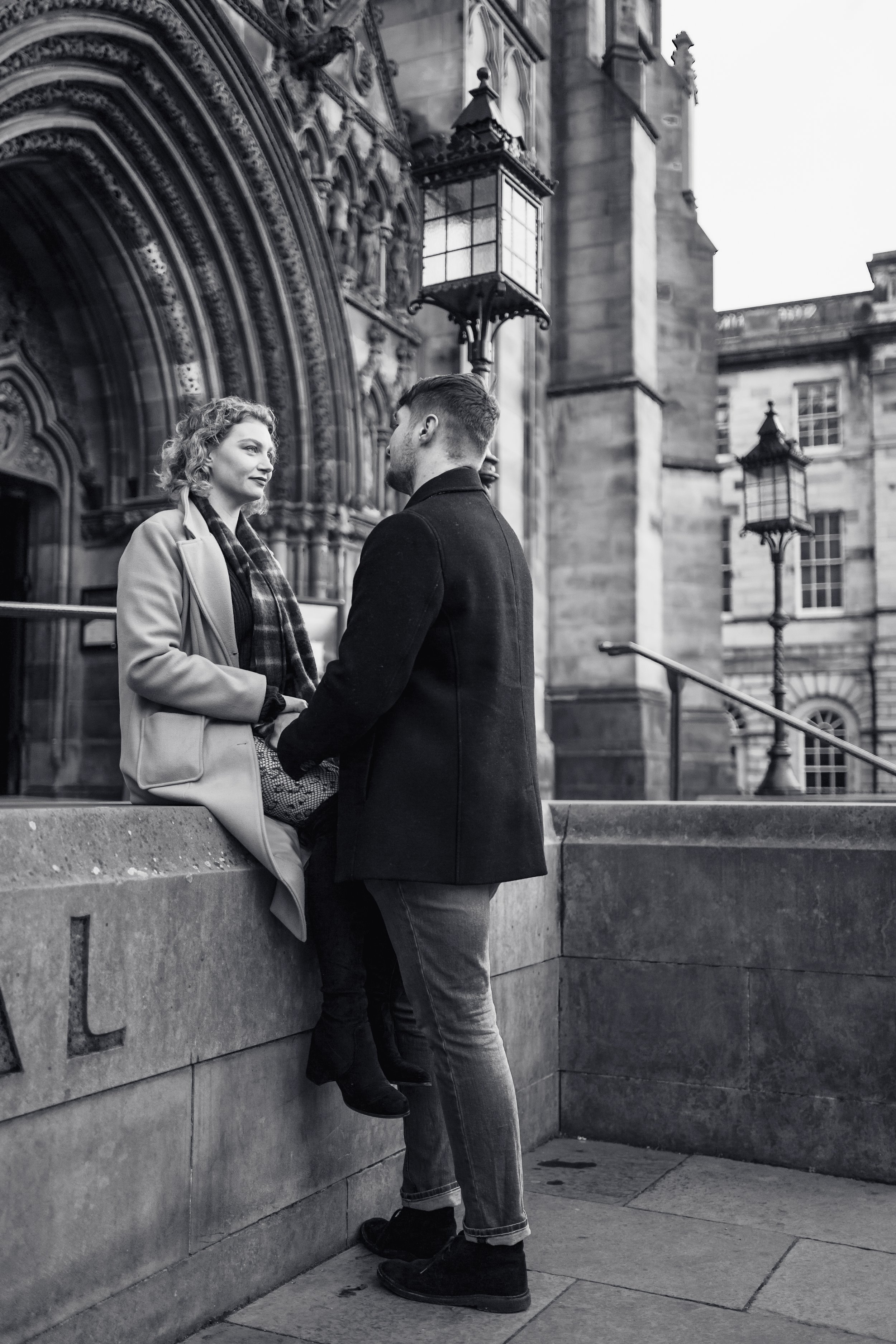 A man and woman are sitting and standing on a stone ledge outside a historic building, engaged in conversation, in black and white photo.