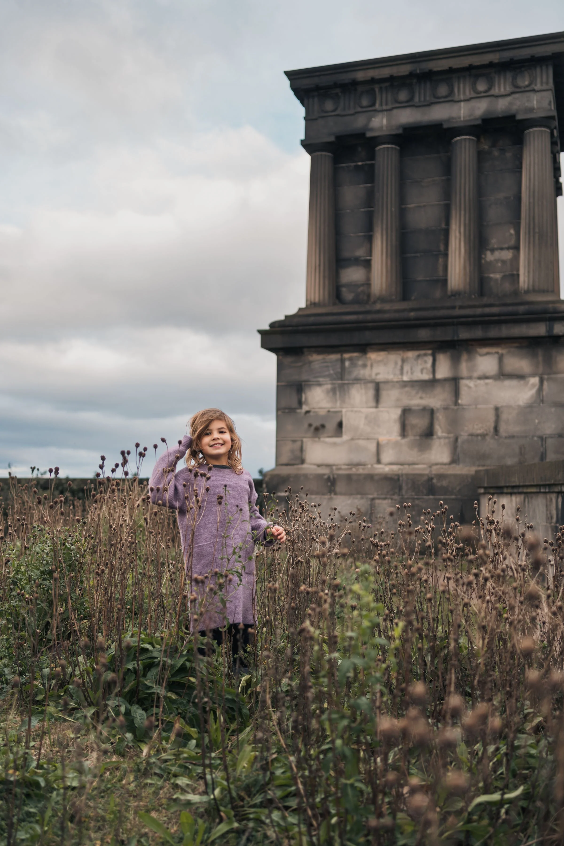 A young girl with wavy hair, wearing a purple sweater and black pants, standing among tall, dry wildflowers near a stone monument with classical columns. The sky is cloudy and overcast.