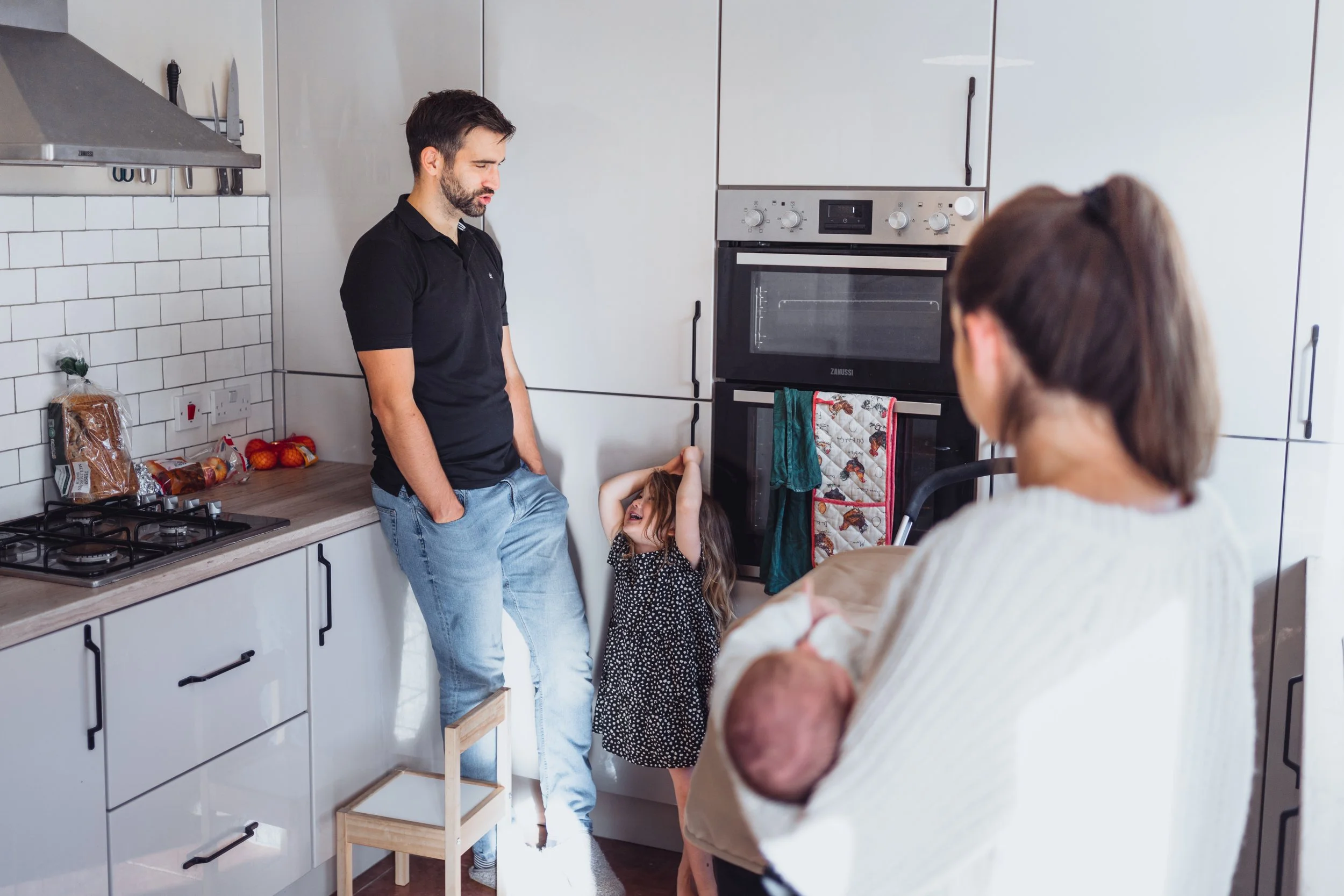 A man leaning against a wall in a kitchen, talking to a young girl with long brown hair, wearing a black and white polka dot dress, who is smiling and holding her head. A woman with brown hair pulled back, holding a baby, is in the foreground facing 