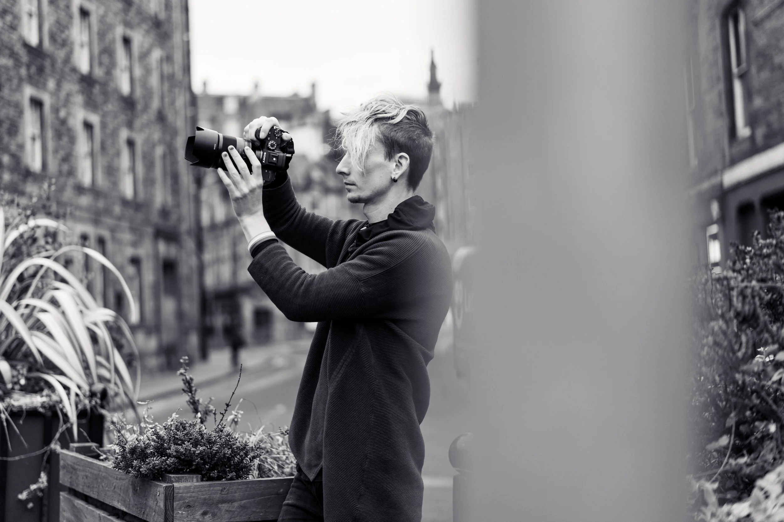A person with short, styled hair taking a photograph with a camera outdoors in an urban area.