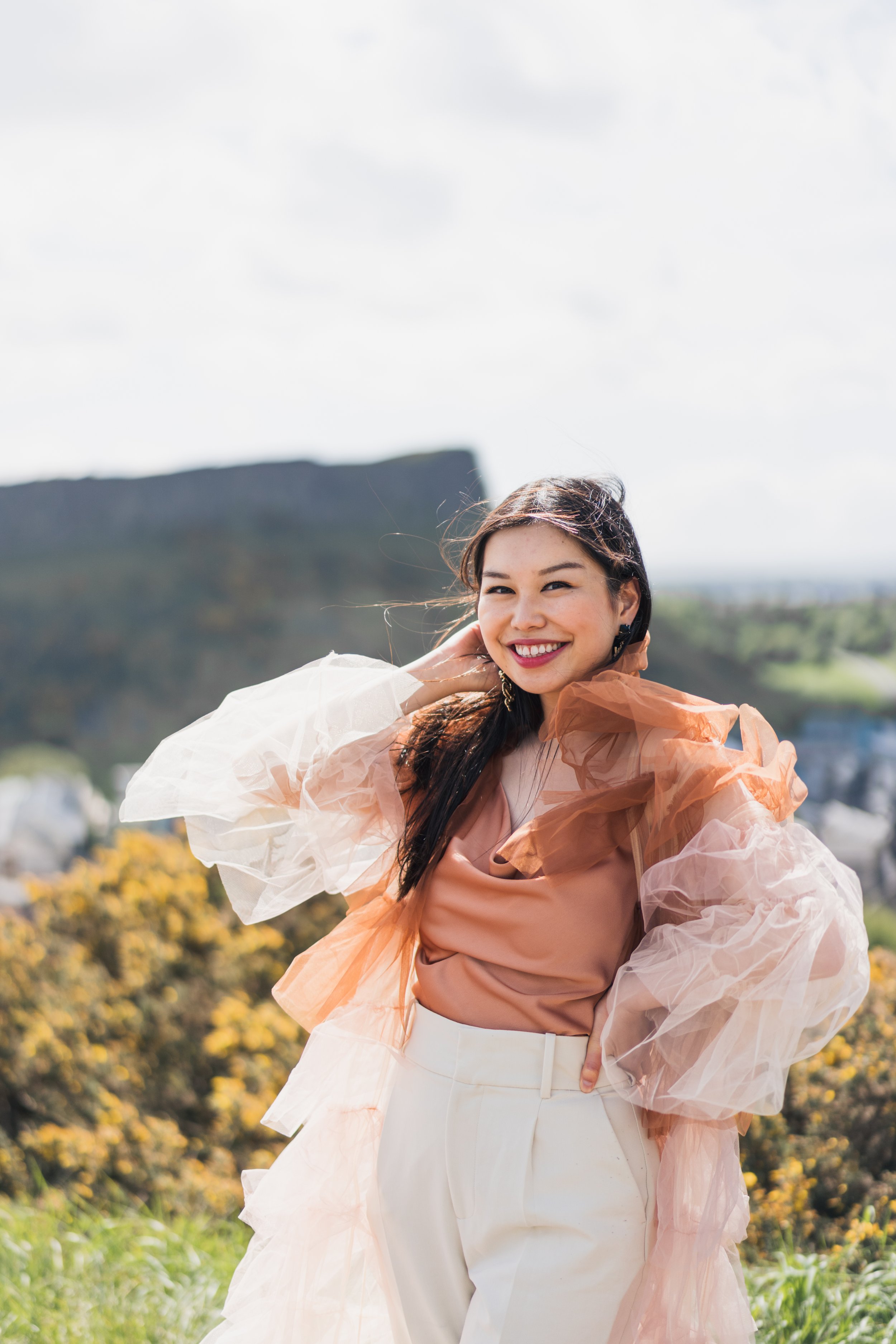 A smiling woman in a peach-colored satin blouse and cream-colored high-waisted pants, with voluminous sheer puff sleeves, standing outdoors with a blurred background of trees, shrubs, and a mountain.
