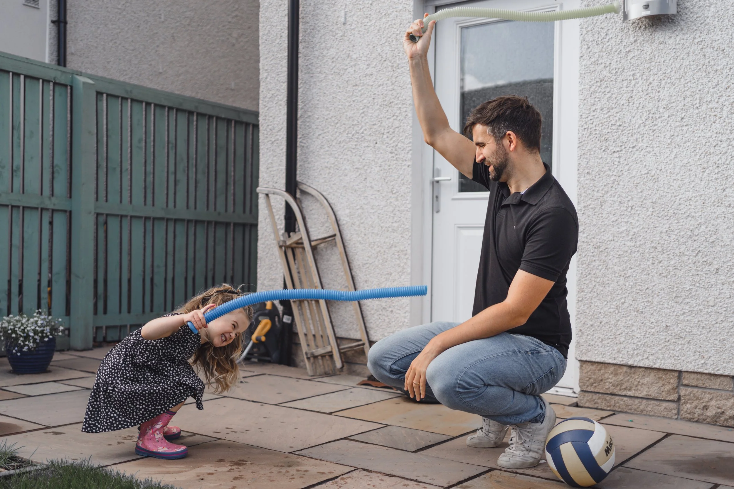 A man and a young girl playing with water outside on a patio. The man is squatting and holding a hose, while the girl is crouching, spraying water with a blue hose, and wearing pink rain boots. A volleyball rests on the ground beside him.