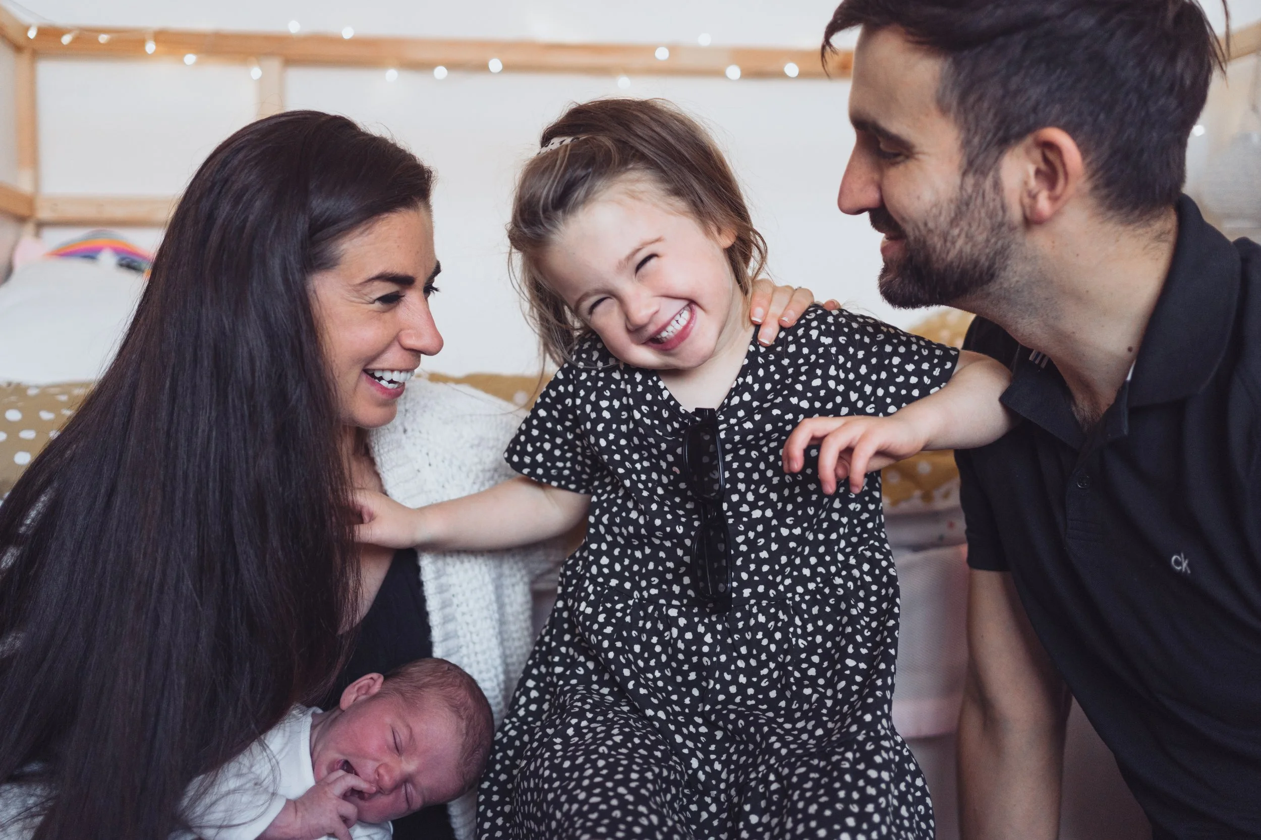 A family of five smiling and interacting indoors, including a woman, a man, two young children, and a newborn baby.