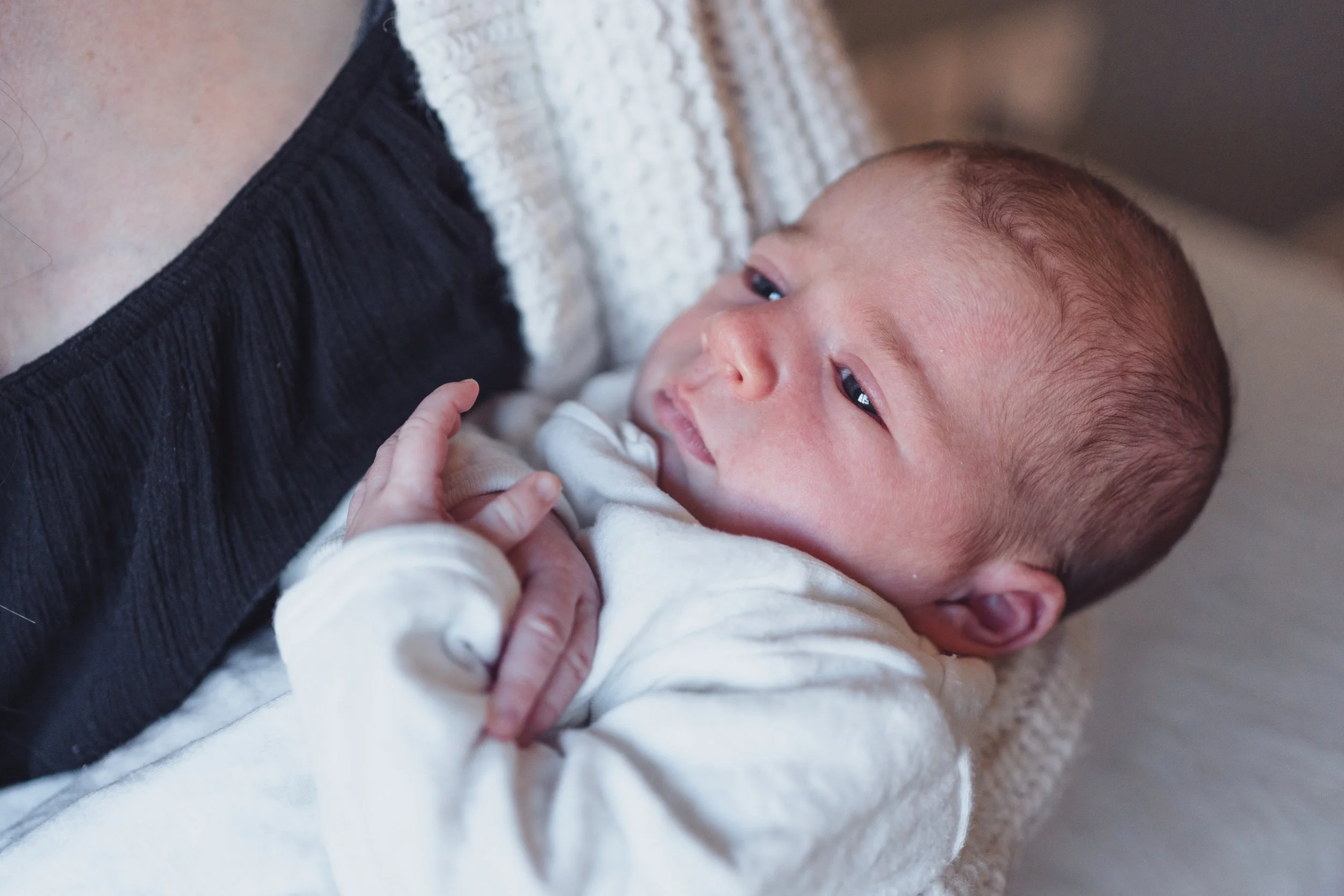 Close-up of a baby holding onto an adult's shirt, lying on a textured blanket, with the baby's face turned slightly towards the camera, showing soft features and short light brown hair.