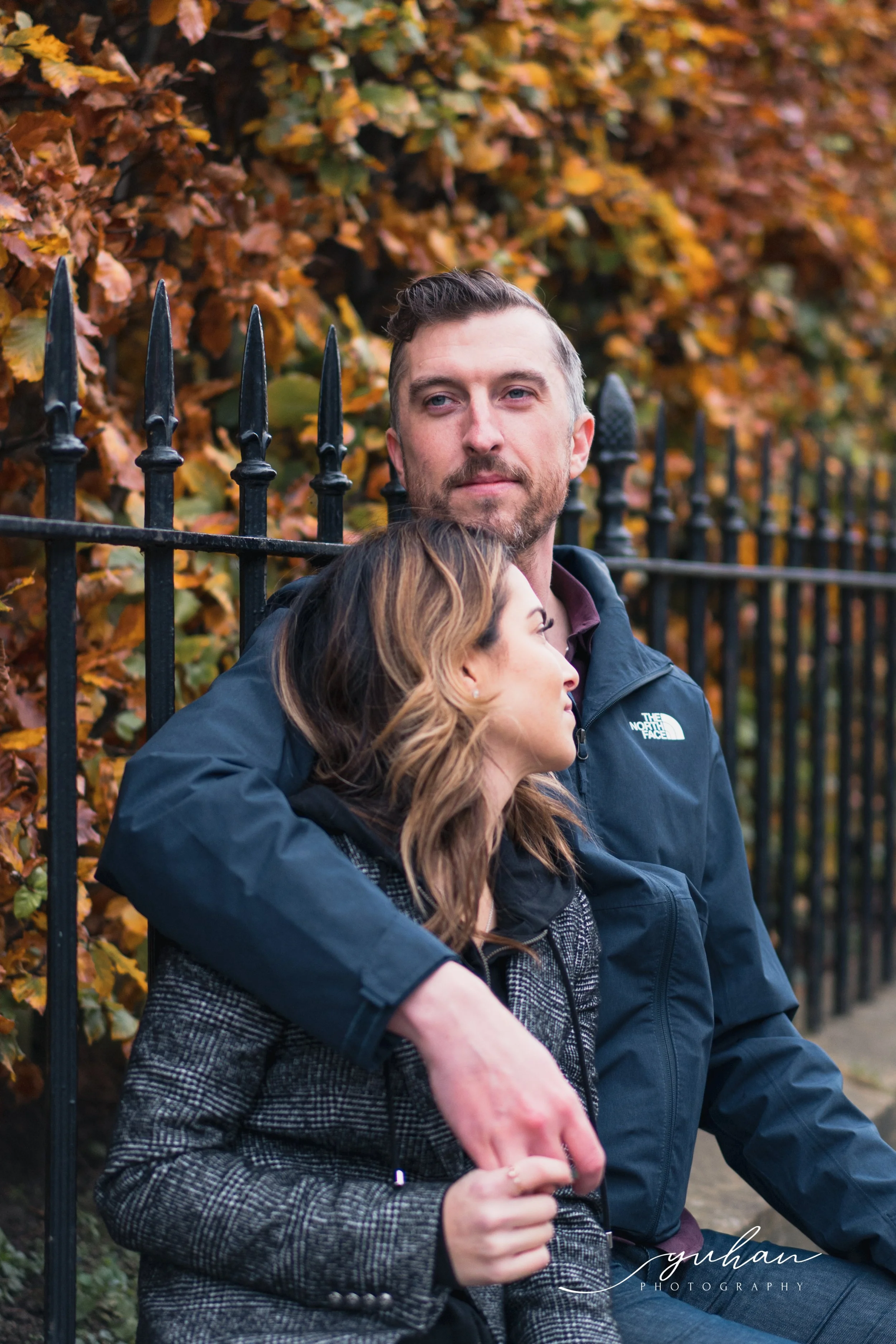A man with a beard and a woman embracing each other, sitting against a black iron fence with autumnal leaves in the background.