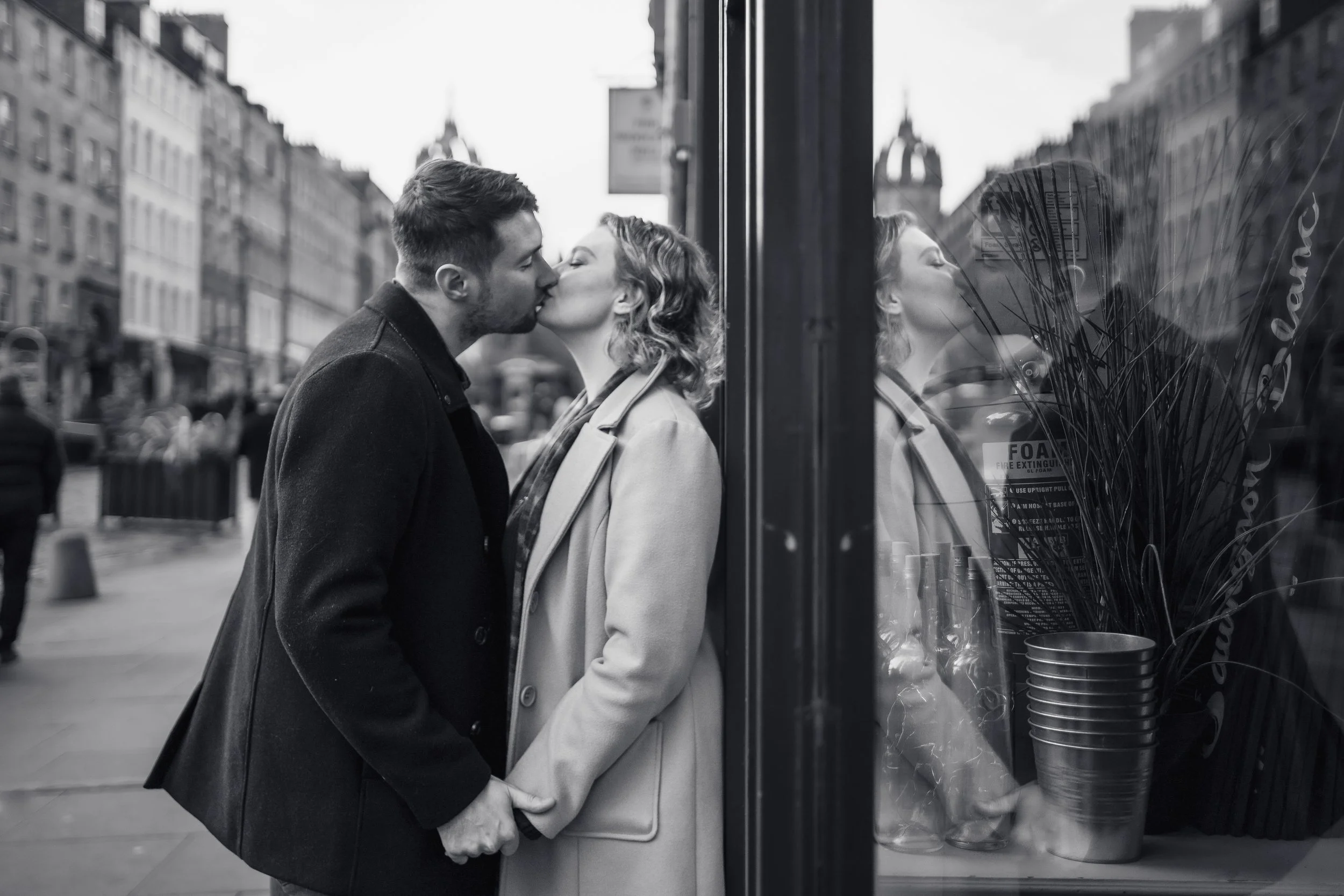 A black-and-white photo of a couple kissing in a city street, with their reflection visible in a storefront window.