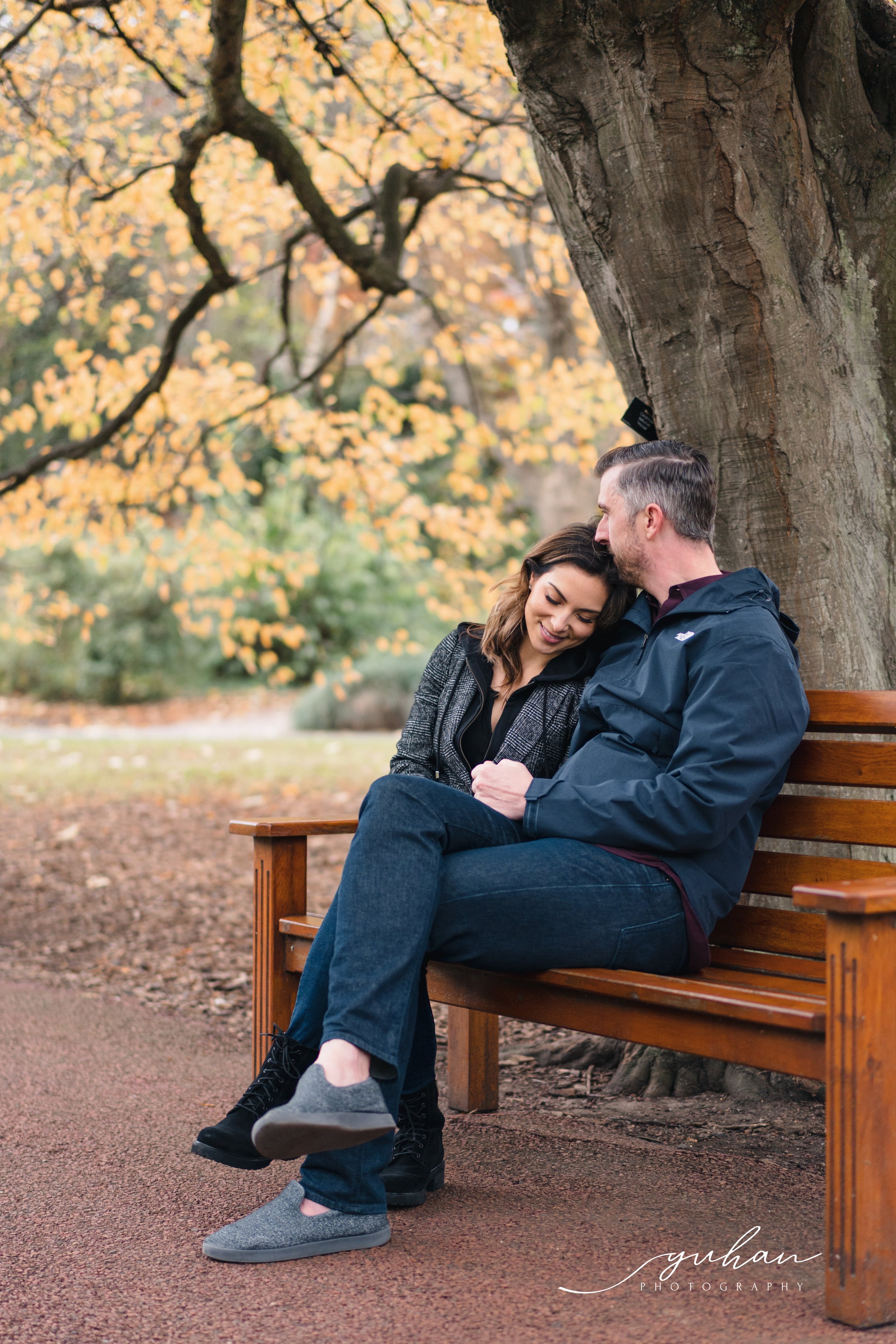 A couple sitting on a wooden park bench, hugging and smiling under a large tree with autumn leaves, surrounded by fallen leaves.