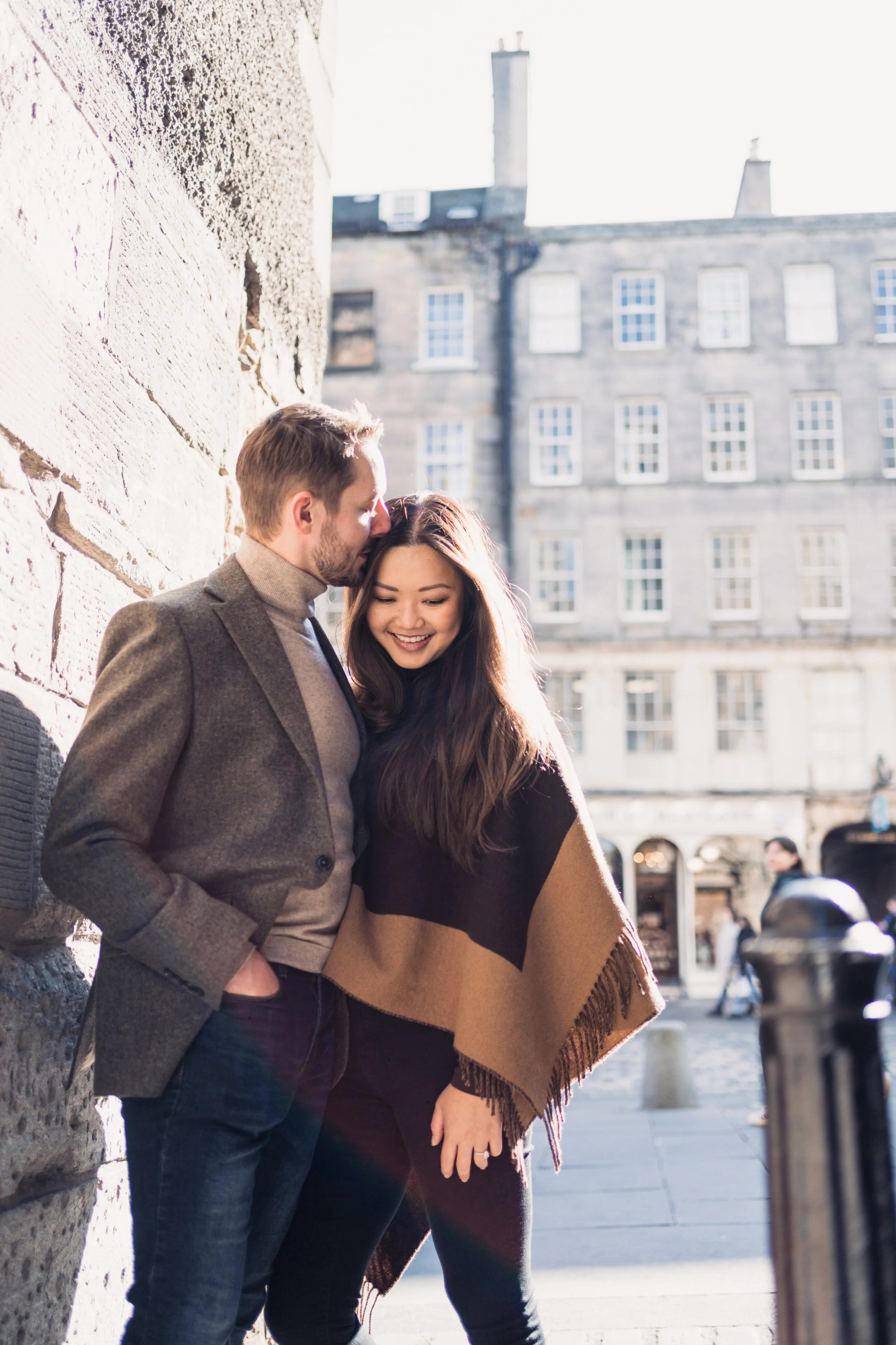 A smiling couple sharing a tender moment on a city street, with the man gently resting his forehead against the woman's, both leaning against a stone wall, with sunlight illuminating their faces.
