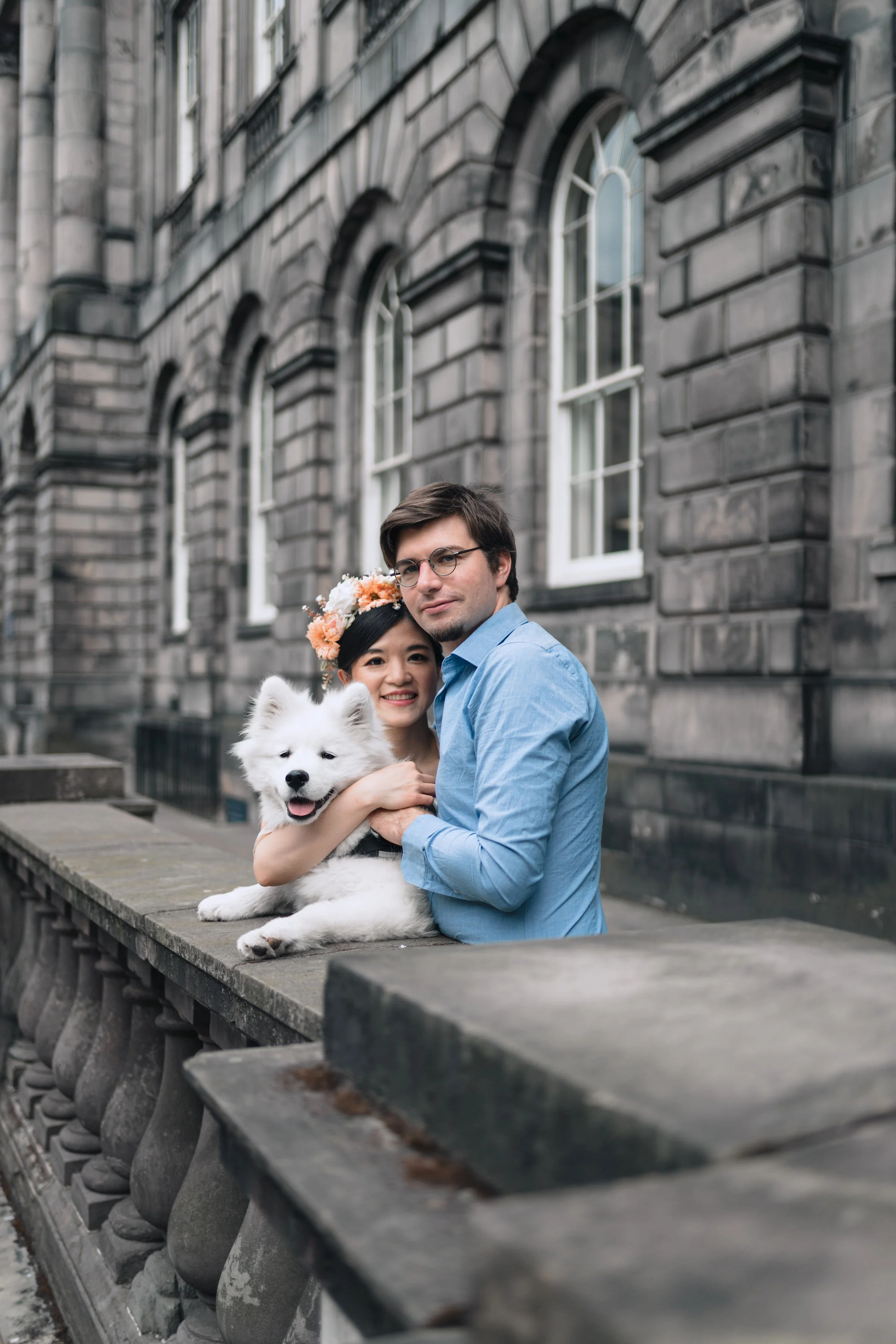 A couple with a dog sitting on a stone railing outside a gray stone building with arched windows.