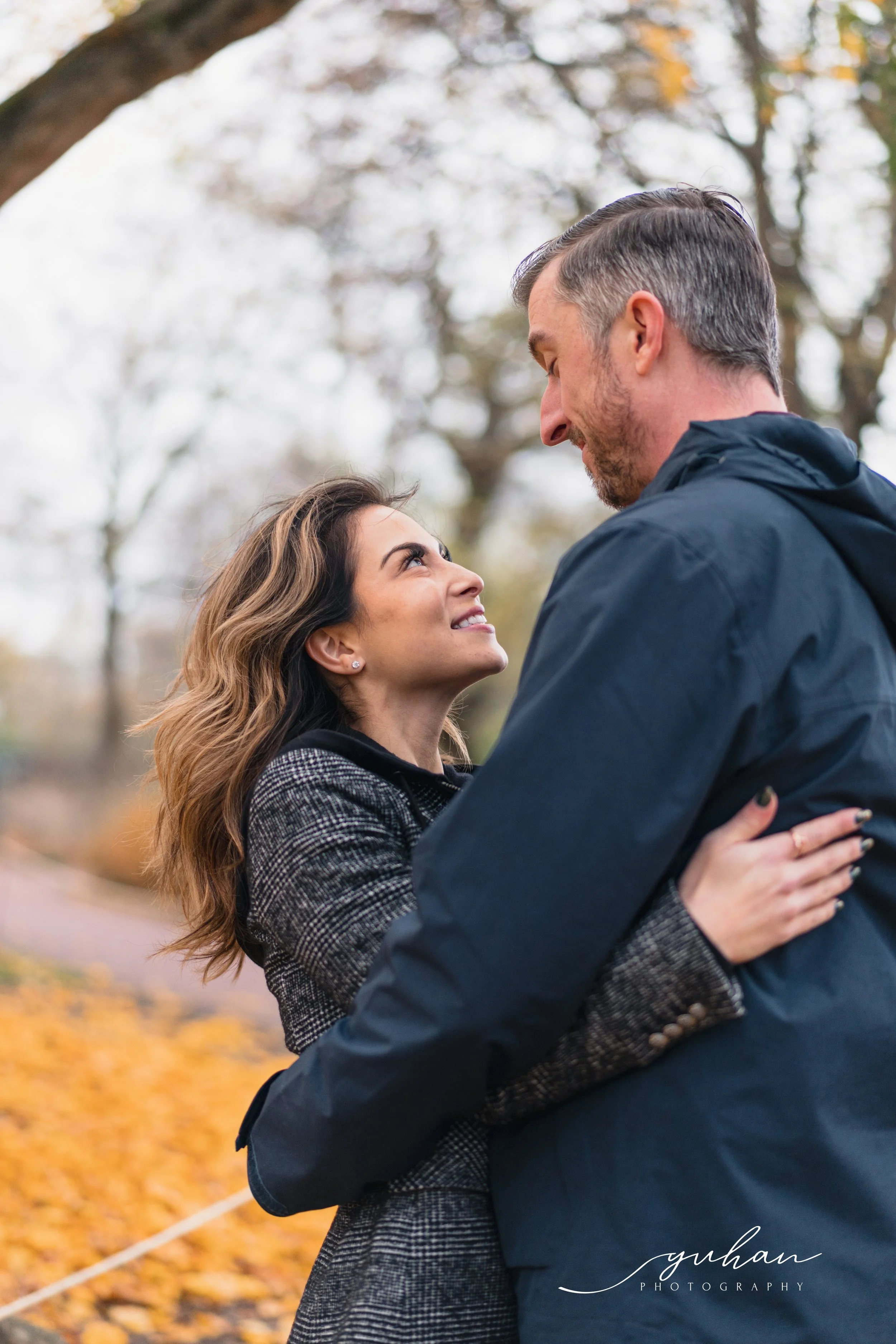 A couple standing outdoors during fall, gazing into each other's eyes, with autumn leaves on the ground and trees in the background.