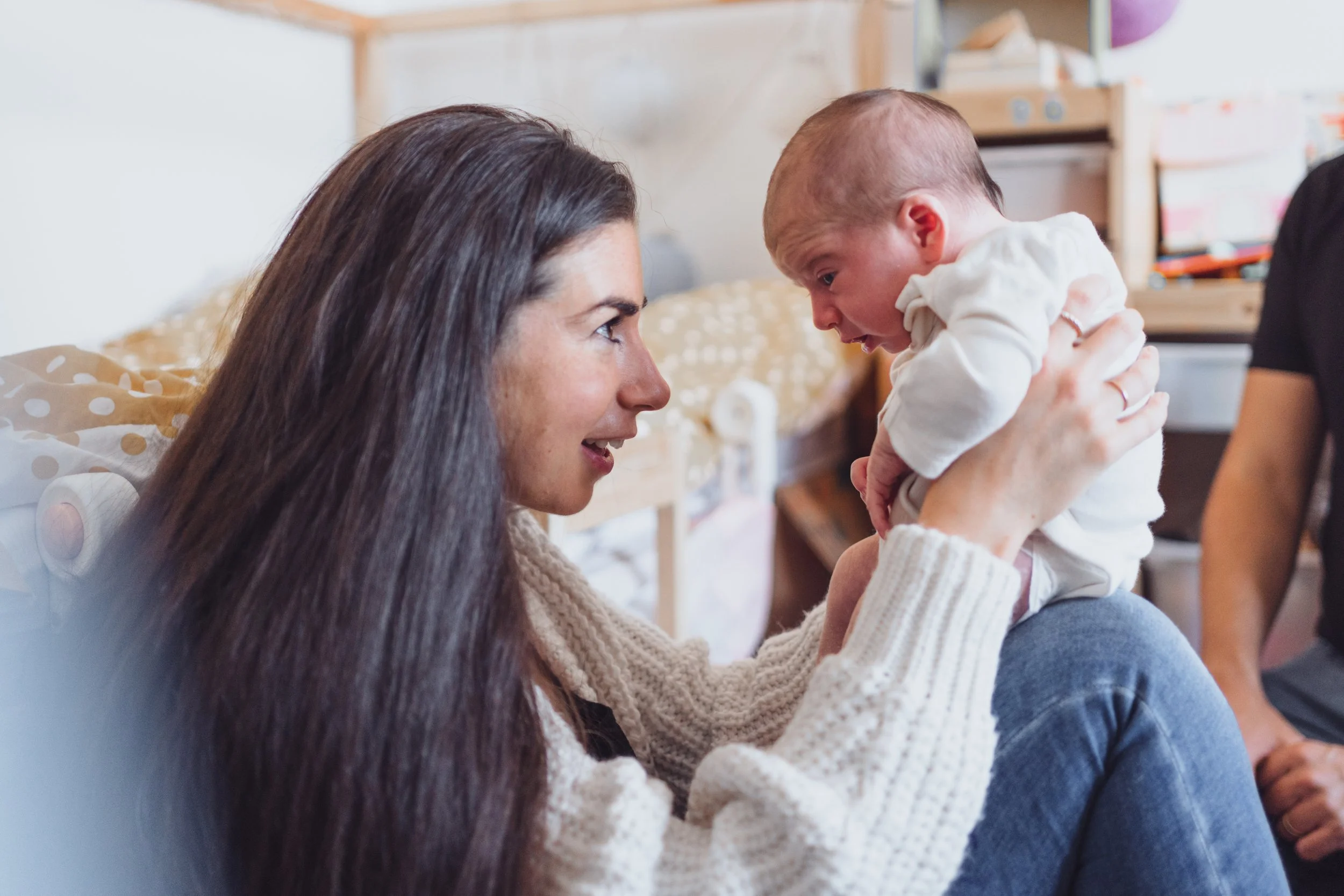 A woman with long dark hair smiling and holding a baby boy who is reaching towards her face, indoors in a cozy room.
