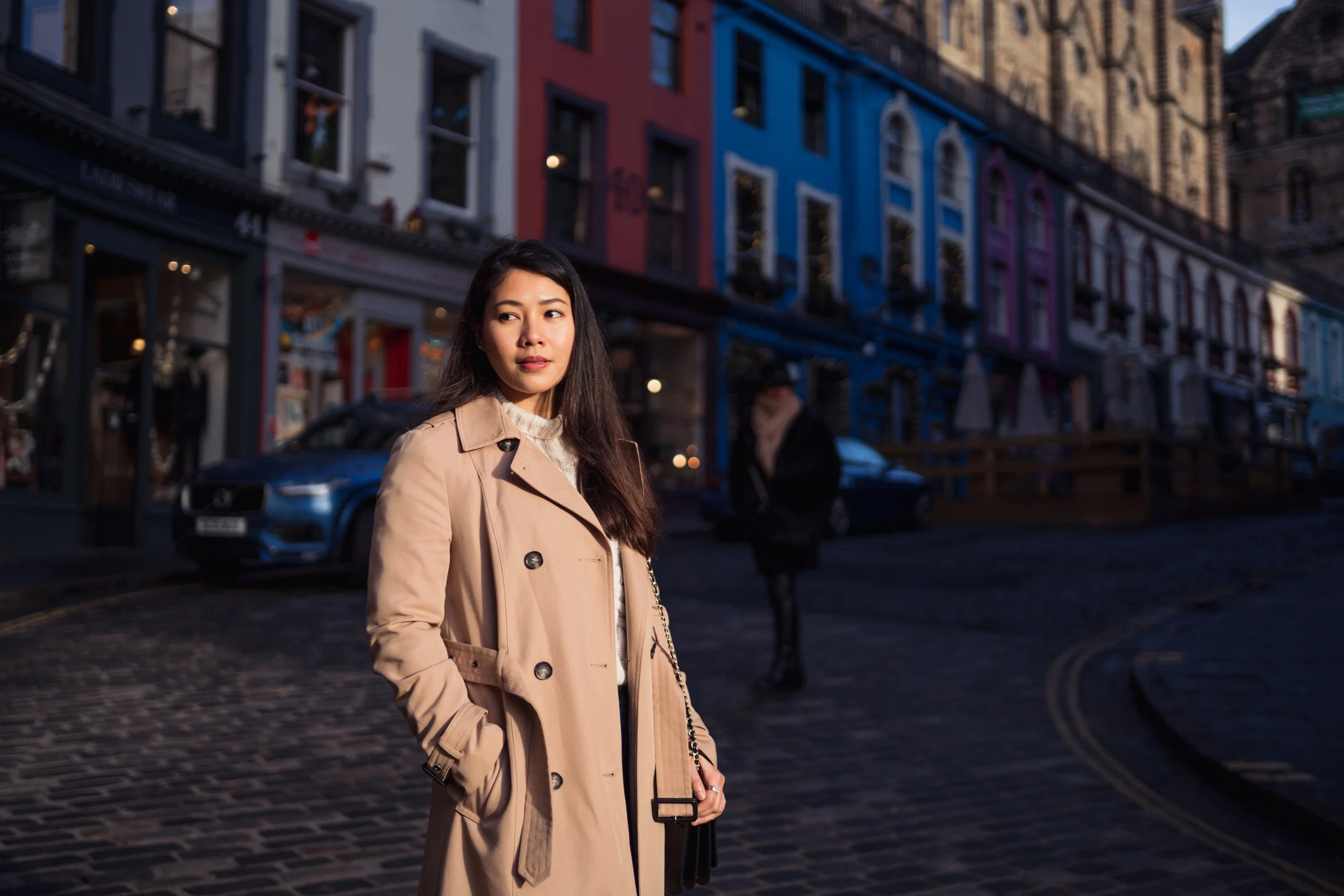A woman in a beige trench coat standing on a cobblestone street in front of colorful buildings with store windows, during the evening or late afternoon.