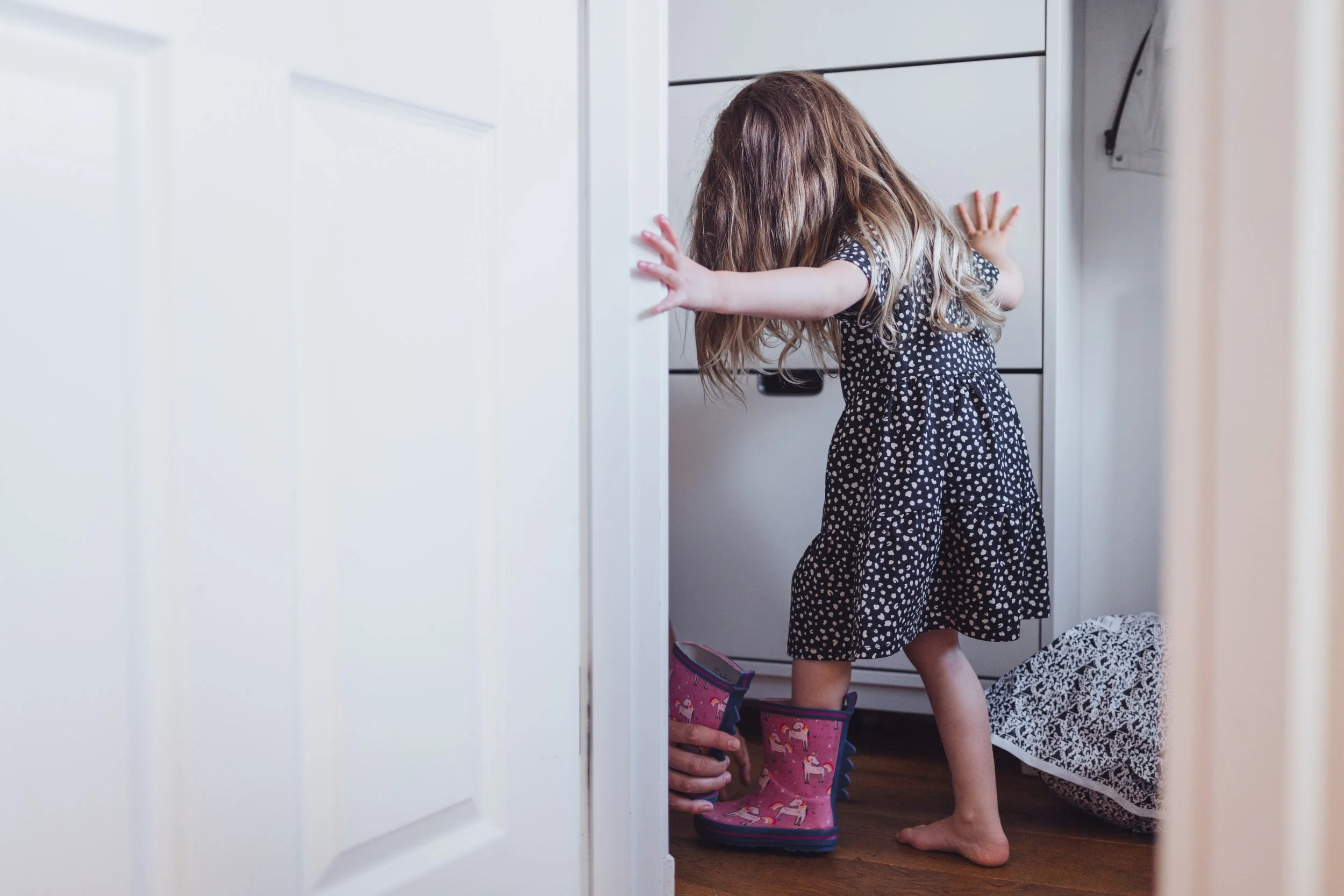 A young girl with long hair wearing a black dress with white polka dots and pink rain boots with unicorns, standing on a wooden floor and reaching into a white cabinet in a room.