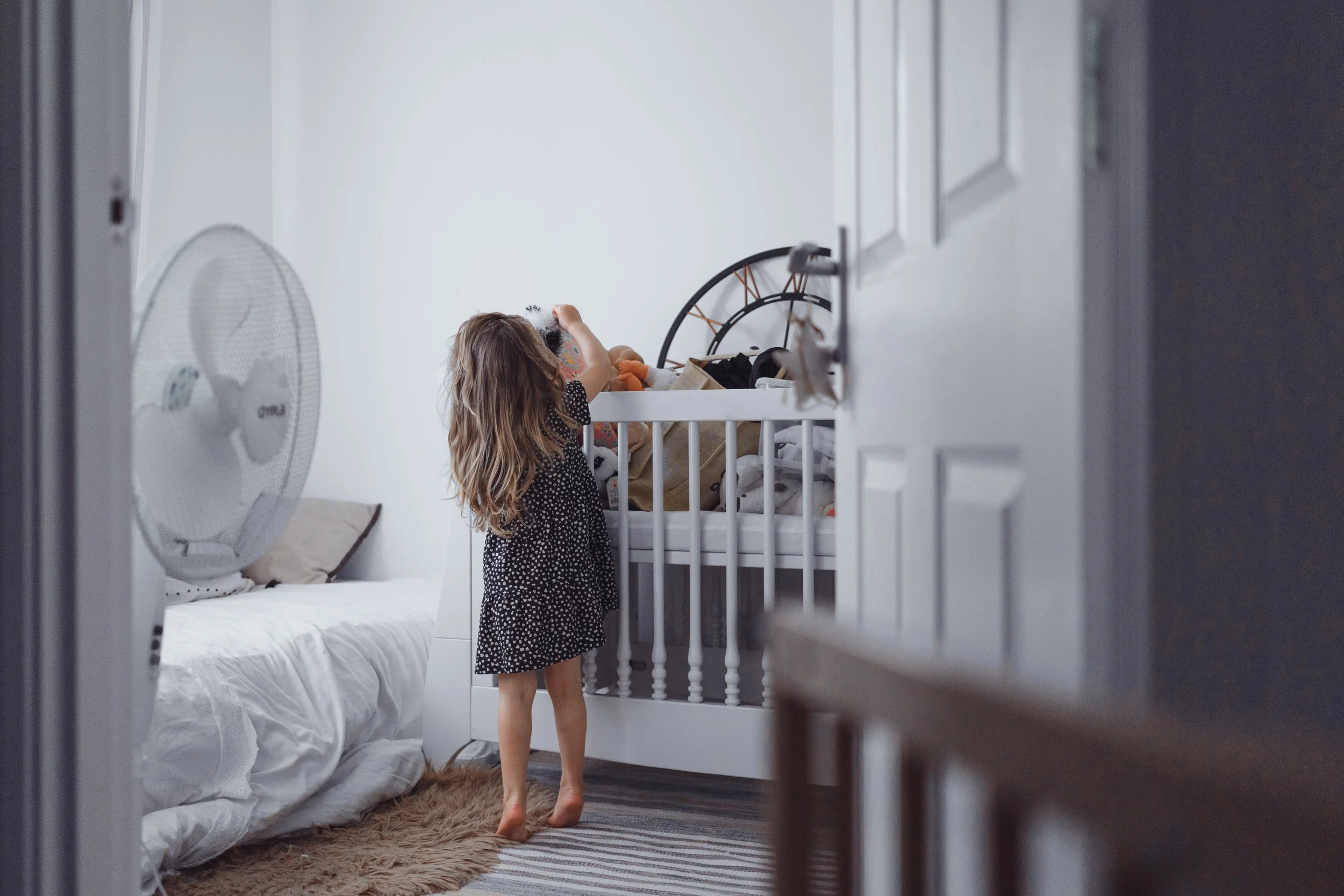 A young girl with long hair in a black polka dot dress is standing barefoot in a bedroom, reaching into a white crib filled with stuffed animals and clothing. A white bed with pillows and a small table with a fan are on the left side of the room.