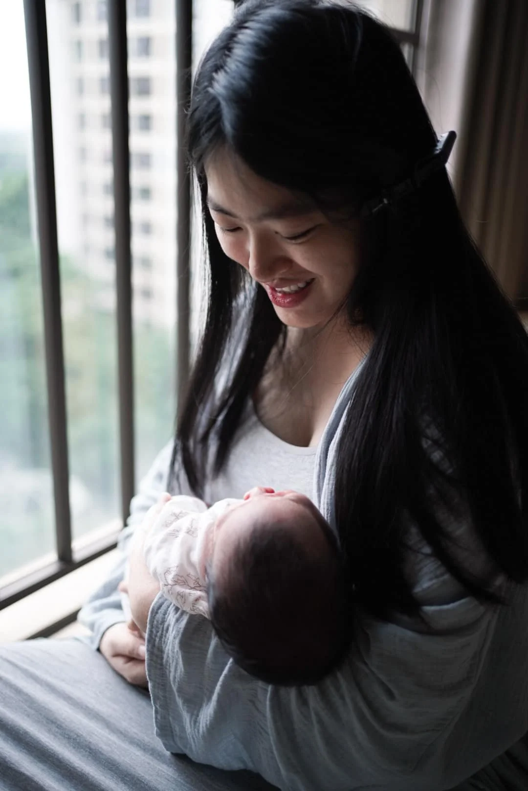 A woman with long dark hair smiling while holding a sleeping baby near a window with city buildings outside.