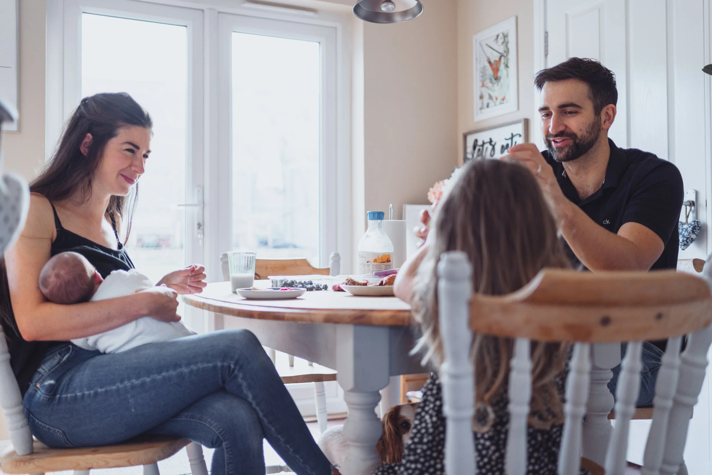 A family enjoying breakfast at a kitchen table with a newborn baby, a young girl, and a dog, near large windows letting in natural light.