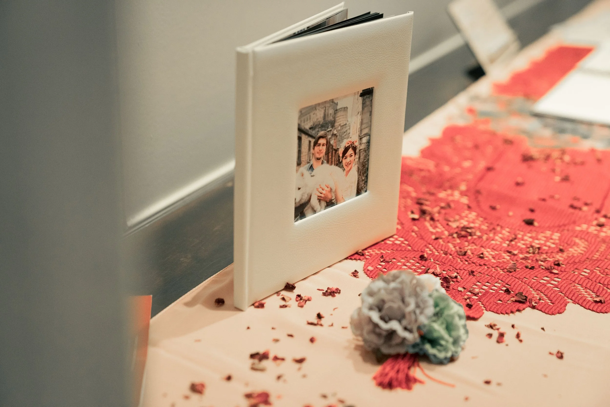 A framed photograph of a smiling couple holding a white dog, placed on a table with a pink and red lace table runner, scattered decorative dried flowers, and a floral arrangement.