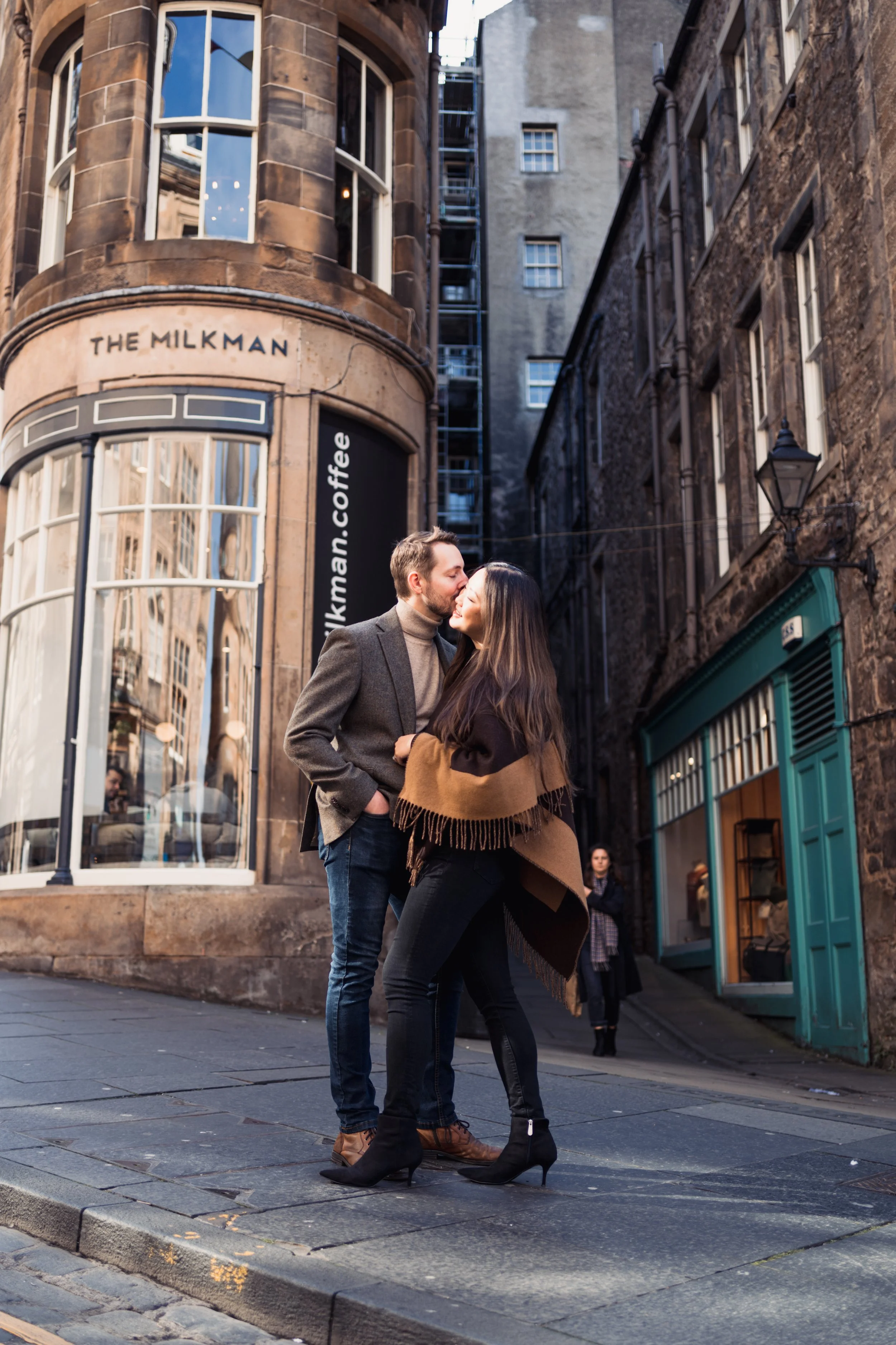 A couple kissing on a city sidewalk in front of a coffee shop named 'The Milkman' with brick buildings surrounding them.