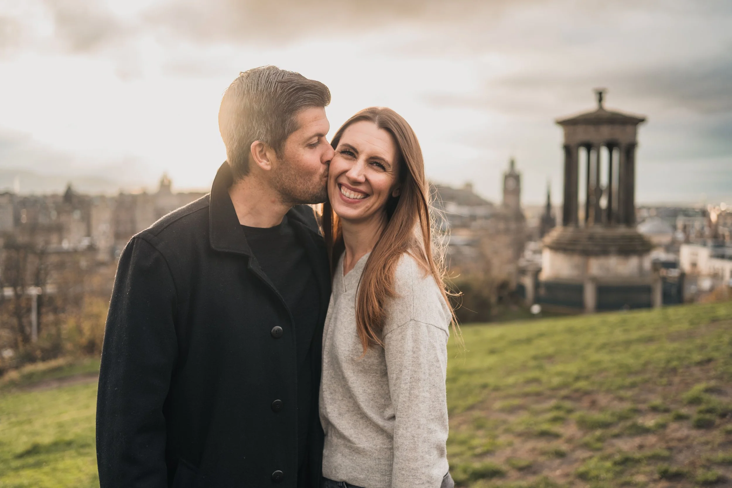 A couple standing outdoors, with the man kissing the woman on the cheek and the woman smiling, in front of a historic cityscape with a classical monument and clock tower in the background.
