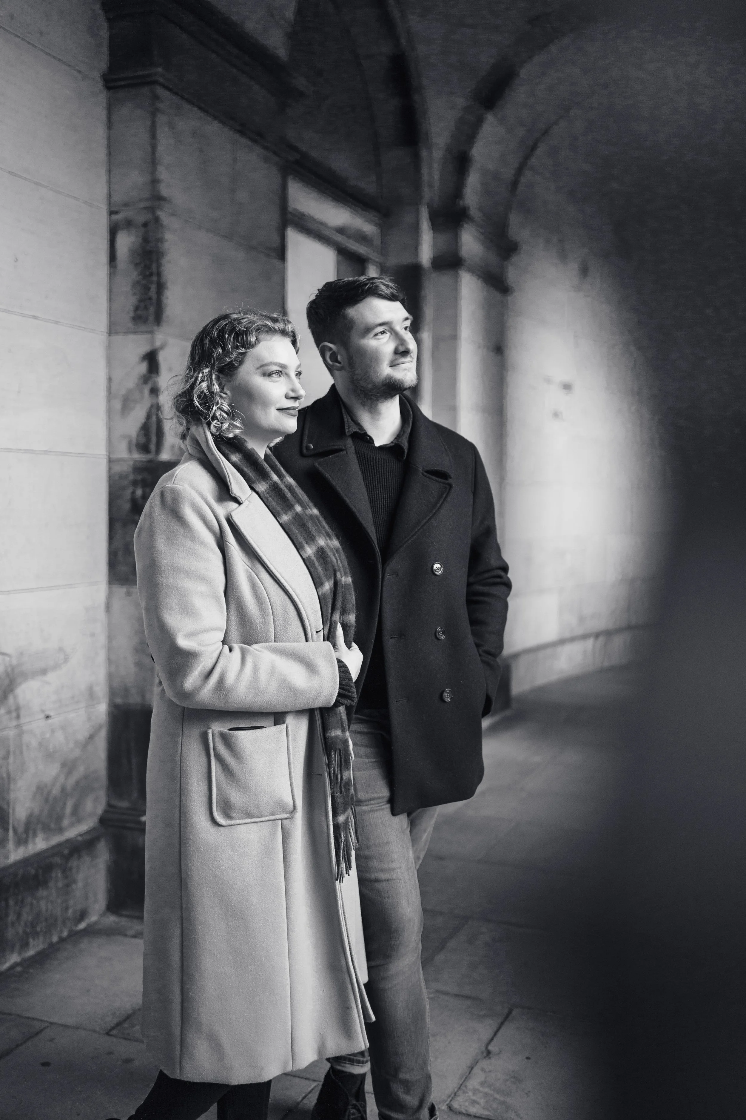 A black-and-white photo of a man and woman standing outside a building, both looking into the distance with calm expressions. The woman has short curly hair and wears a coat with a scarf, and the man has short hair and a beard, wearing a coat with hi