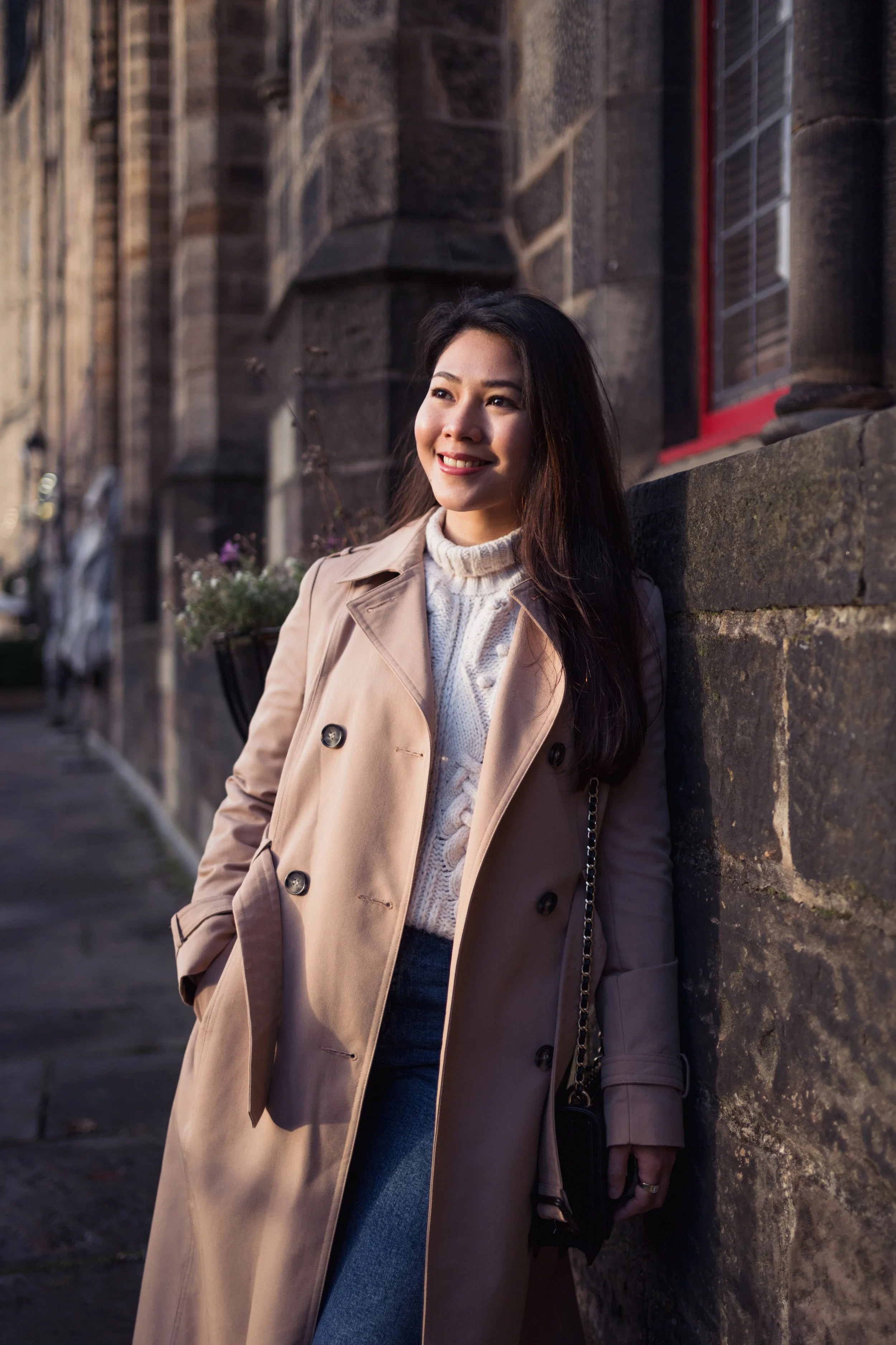A woman leaning against a stone wall on a city sidewalk, smiling and looking to her right. She is wearing a beige trench coat over a white cable-knit sweater, with dark jeans, and carrying a small black purse with a chain strap.