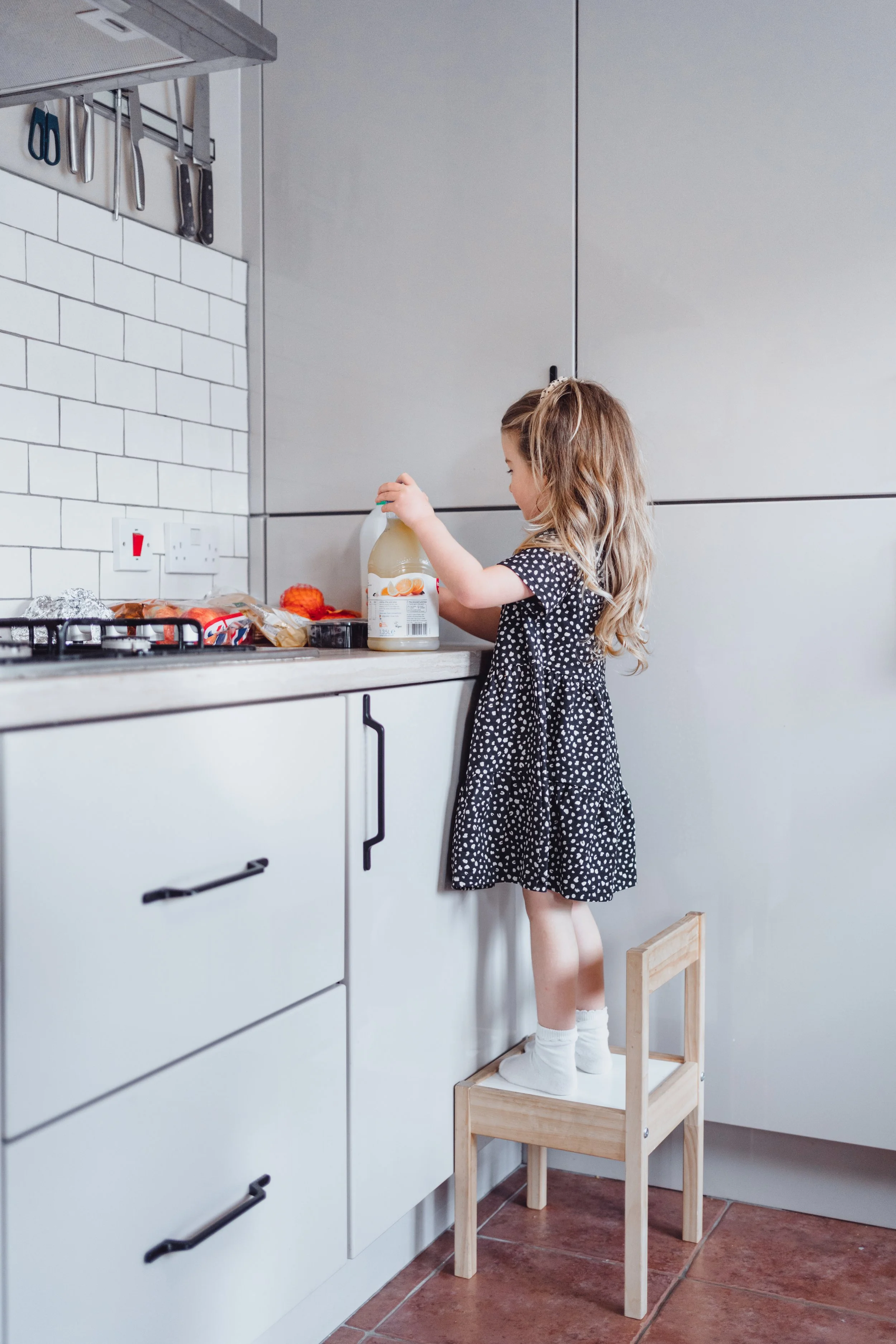 A young girl in a black and white polka dot dress standing on a small wooden chair to reach the kitchen counter, holding a bottle of juice with other food items on the counter.