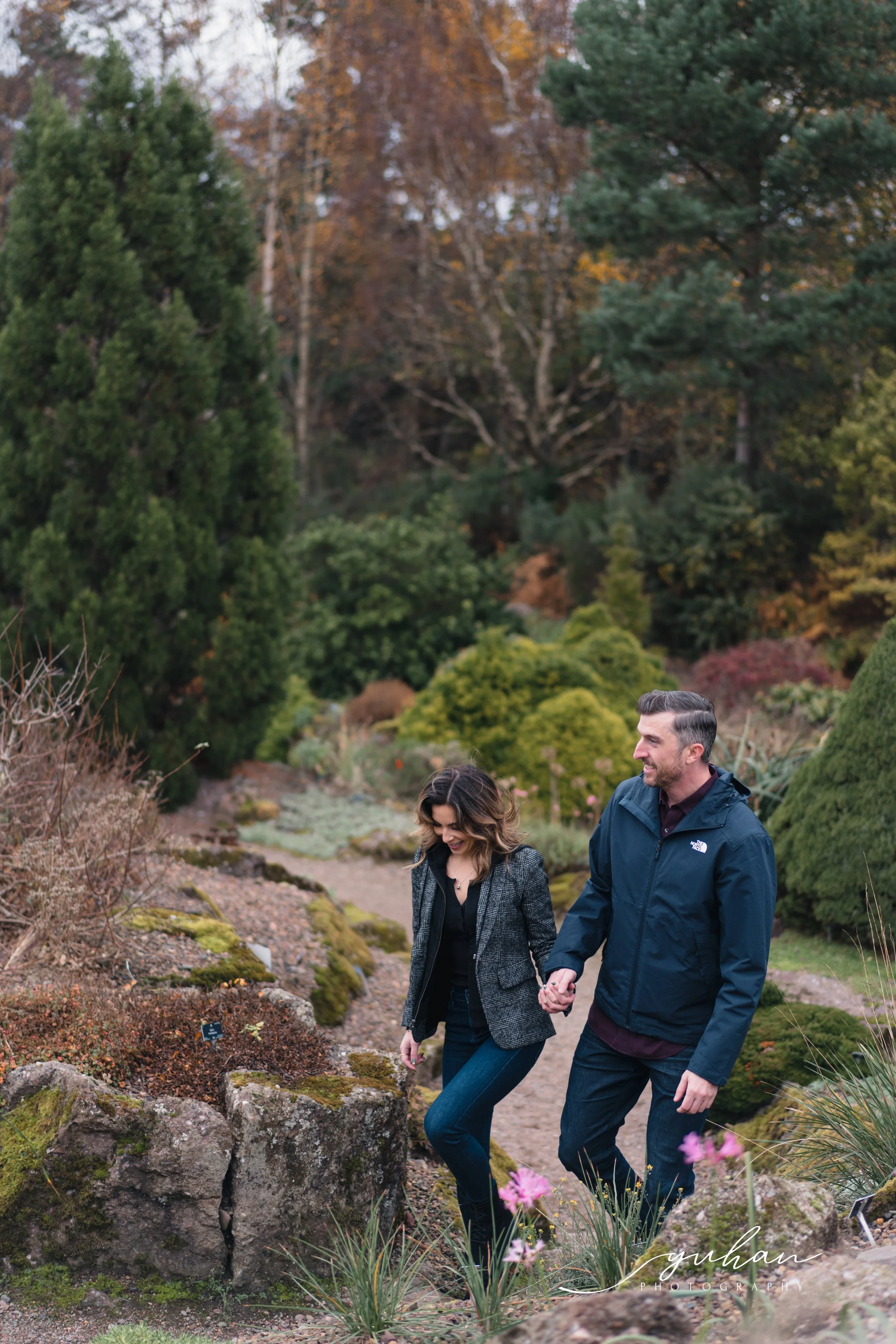 A couple walking hand in hand through a lush, manicured garden with various trees and plants during autumn.