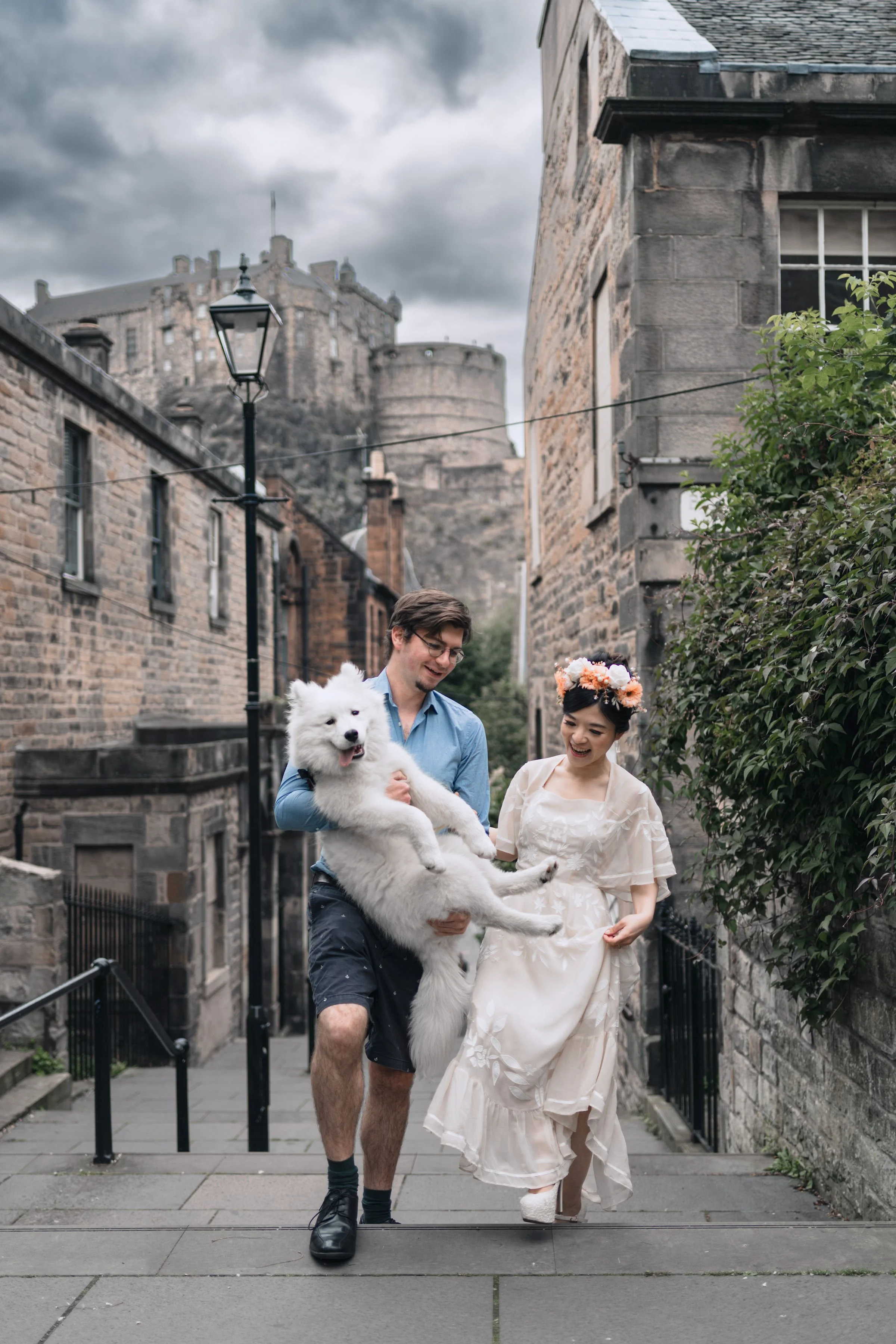 A couple and their fluffy white dog walking up a cobblestone street with historic stone buildings and a castle on a hill in the background. The woman is in a white dress and floral crown, the man is in a blue shirt and shorts, and they appear happy.