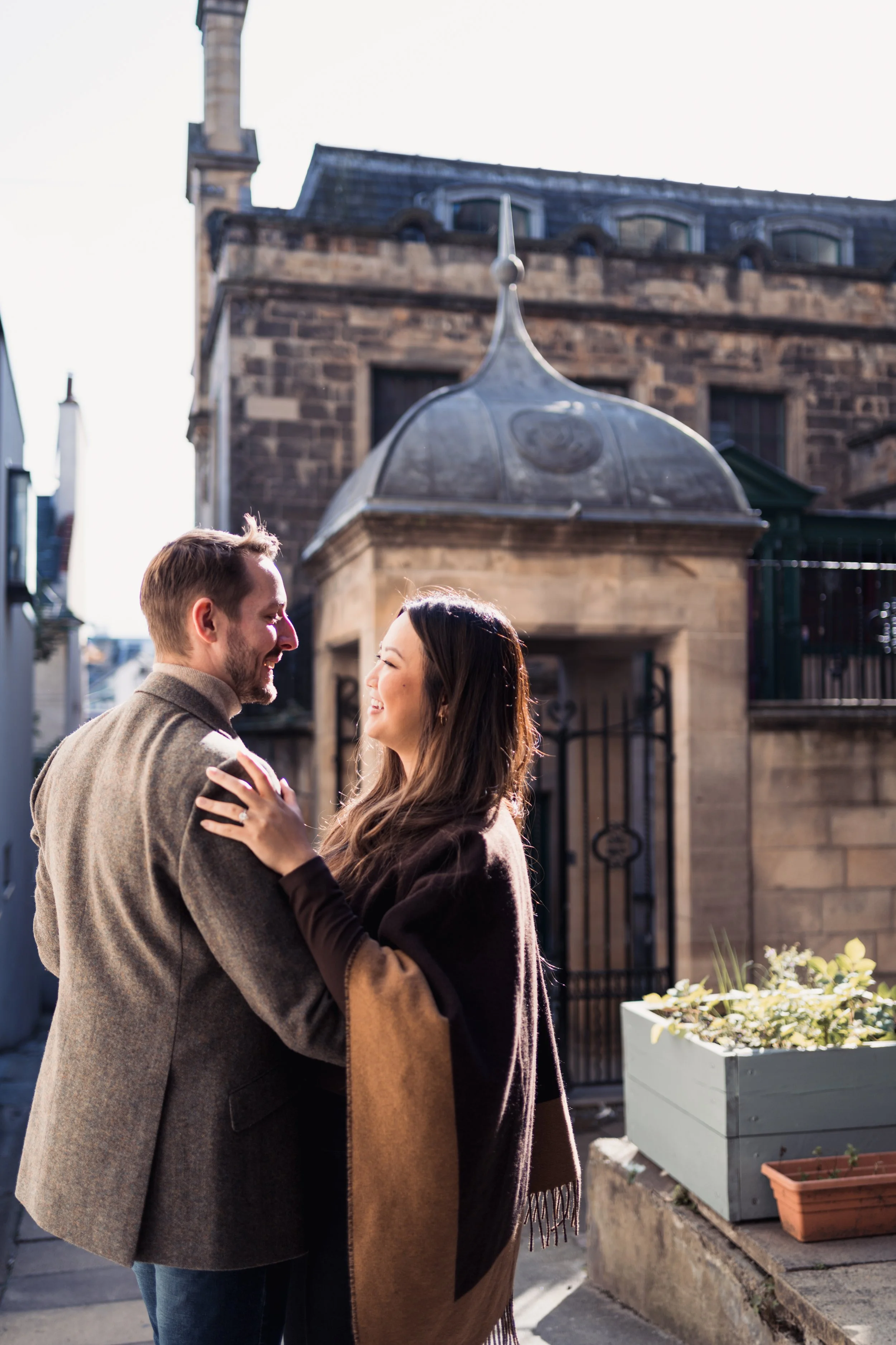 A couple smiling and embracing outdoors in front of an old stone building with a clock tower, during daytime.