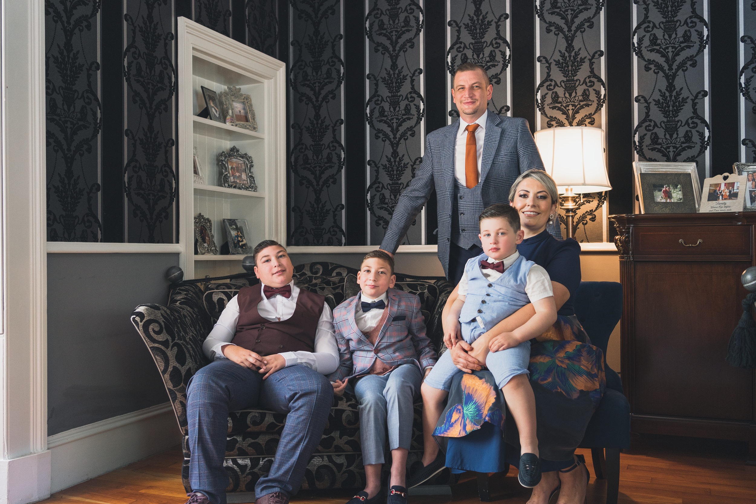 A family of five posing for a photo in a stylish living room. The father is standing behind the sofa, wearing a gray checked suit with an orange tie. The mother is sitting on the sofa, wearing a dark blue dress, with three young boys sitting and stan