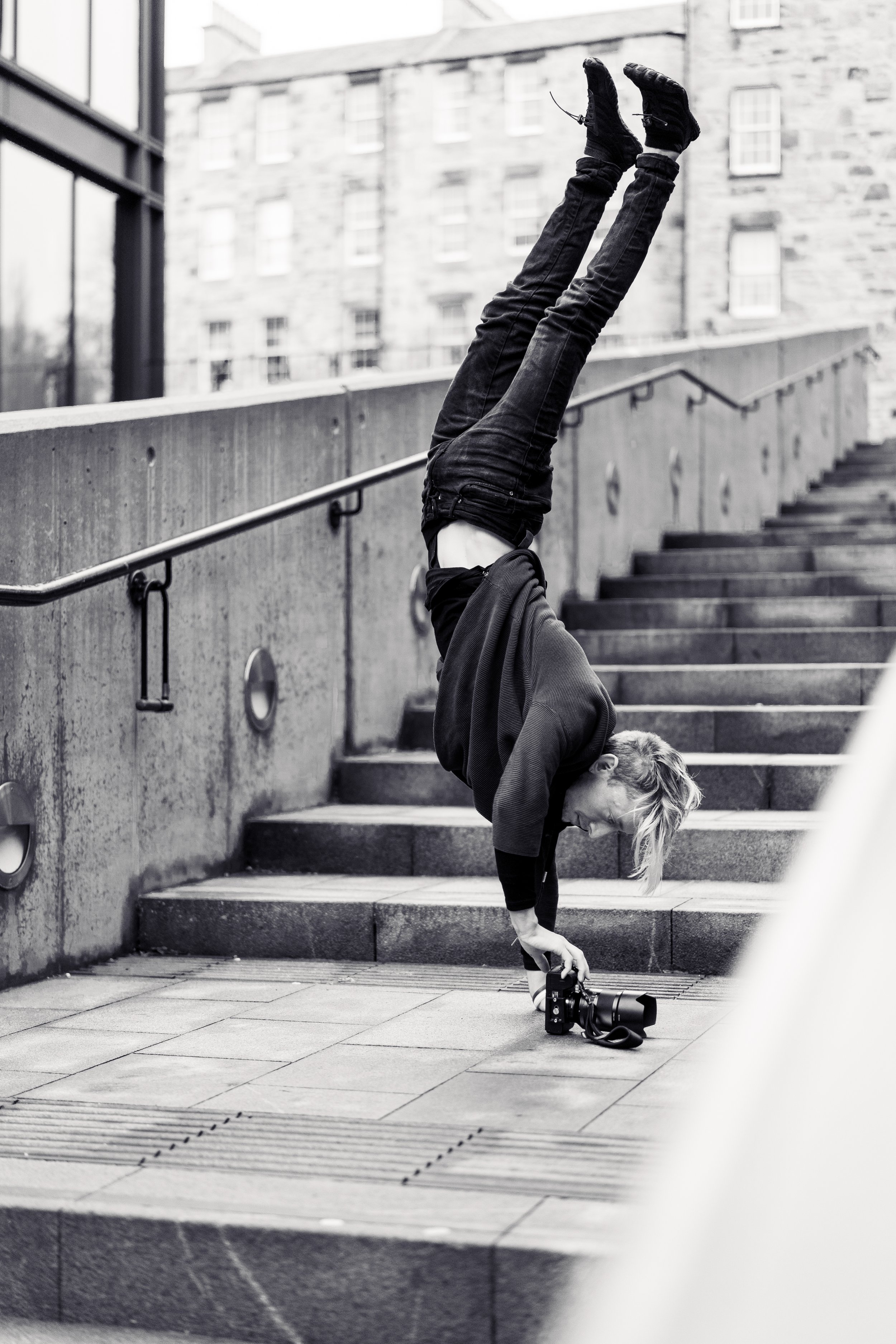 A woman doing a handstand on stairs outdoors, holding a camera with one hand, in black and white.