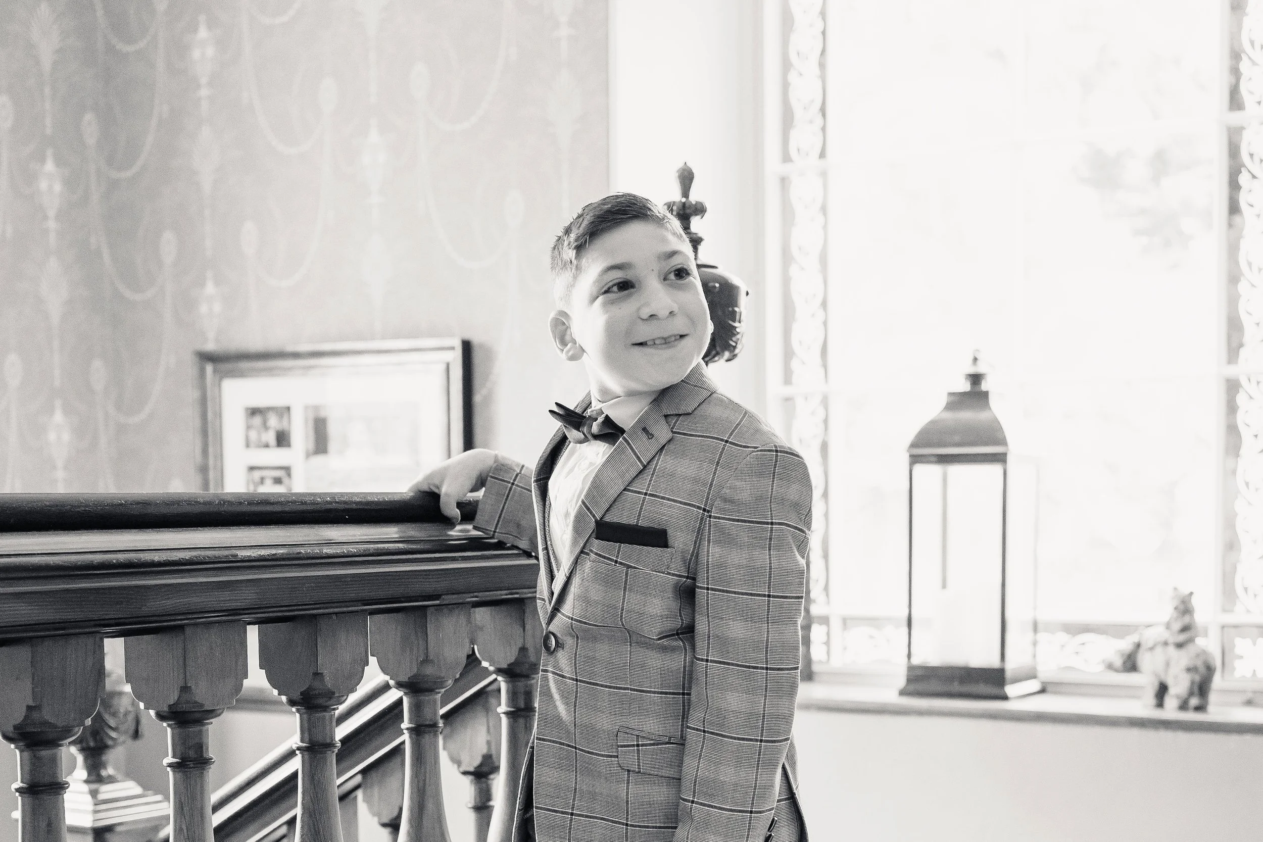 A young boy in a plaid suit with a bowtie smiling, standing indoors next to a wooden railing.