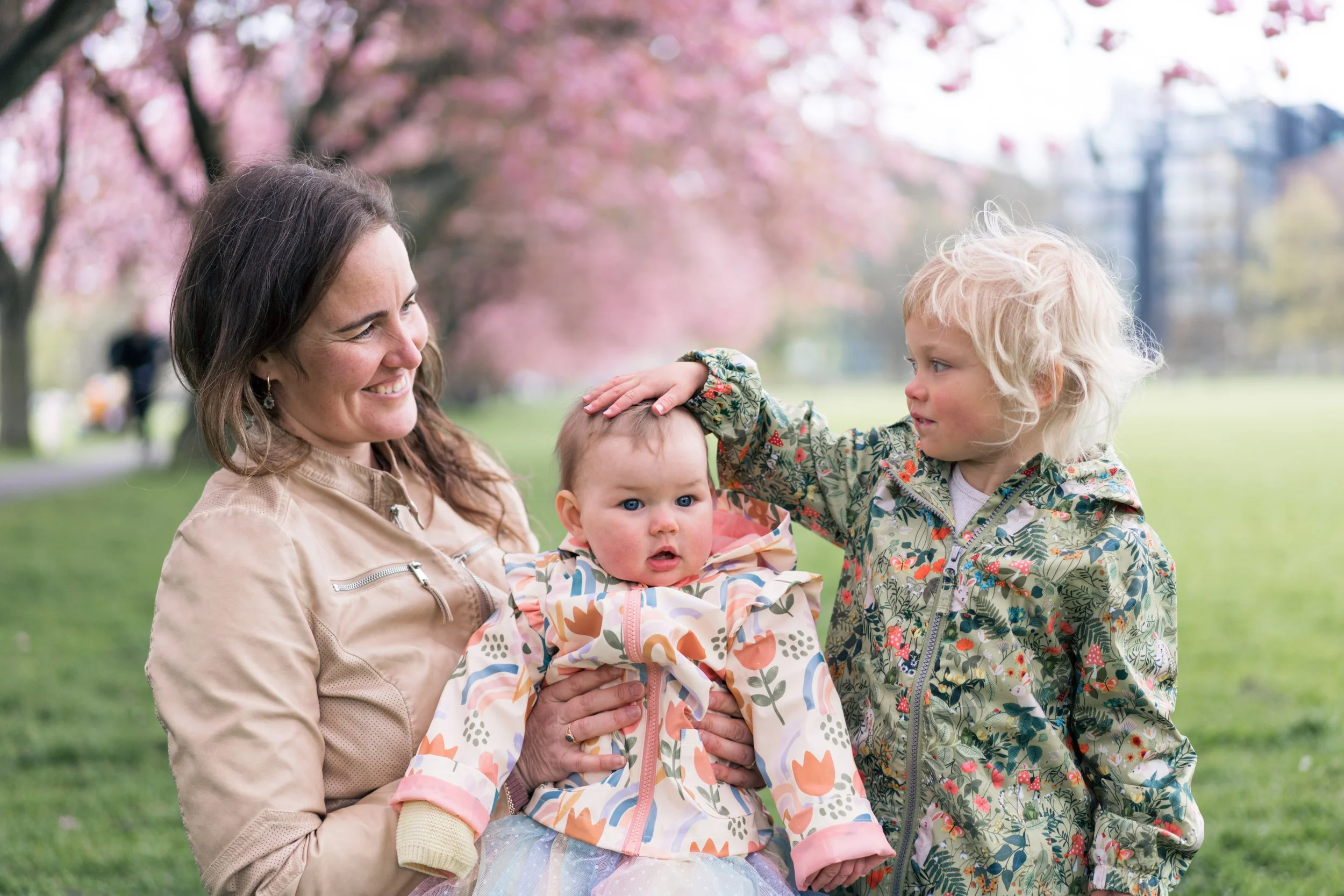 A woman holding a baby is smiling while a young girl gently pats the baby's head in a park with pink blossoming trees.