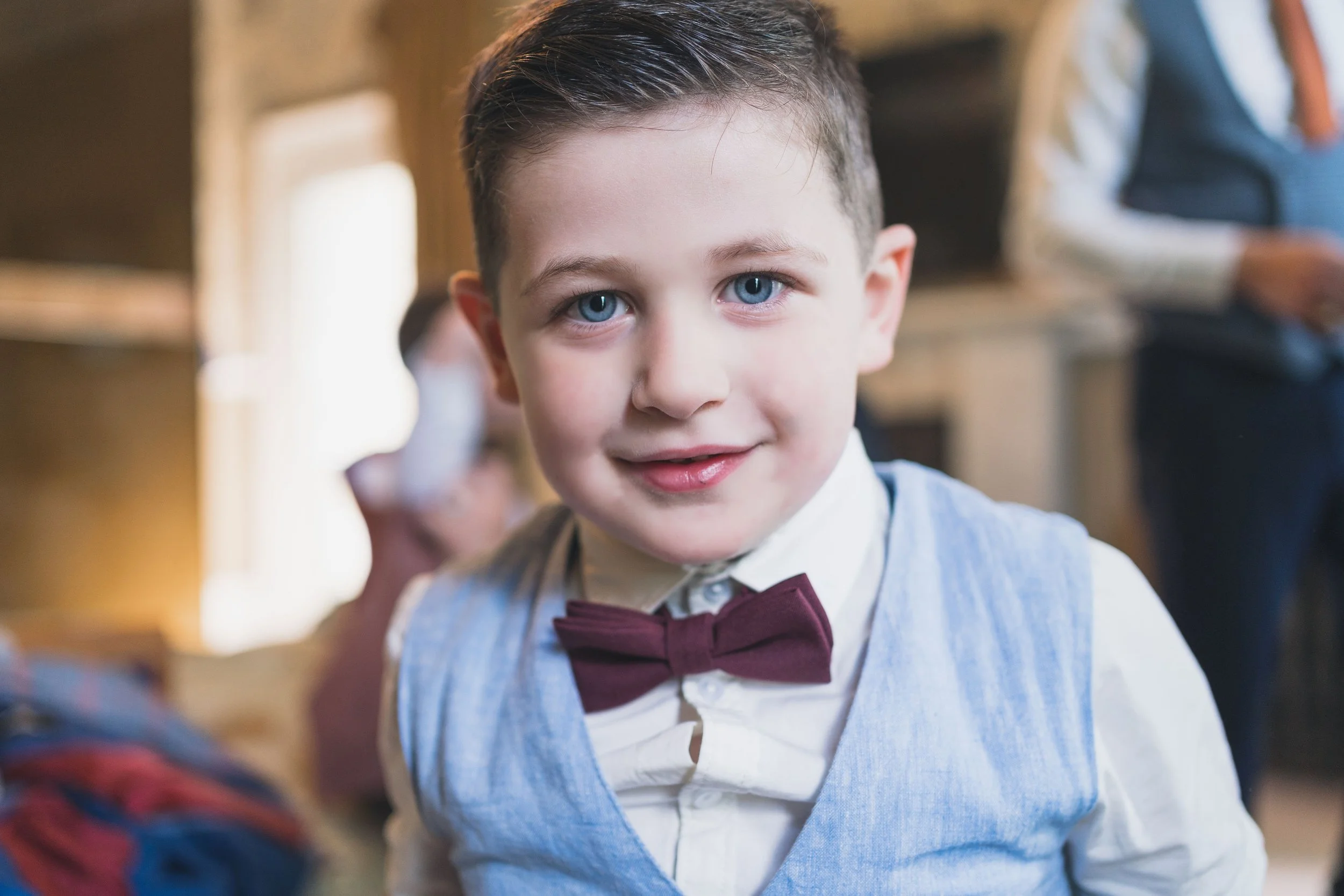 A young boy with blue eyes and brown hair, dressed in a white shirt, light blue vest, and a burgundy bow tie, smiling and looking at the camera in a classroom setting.
