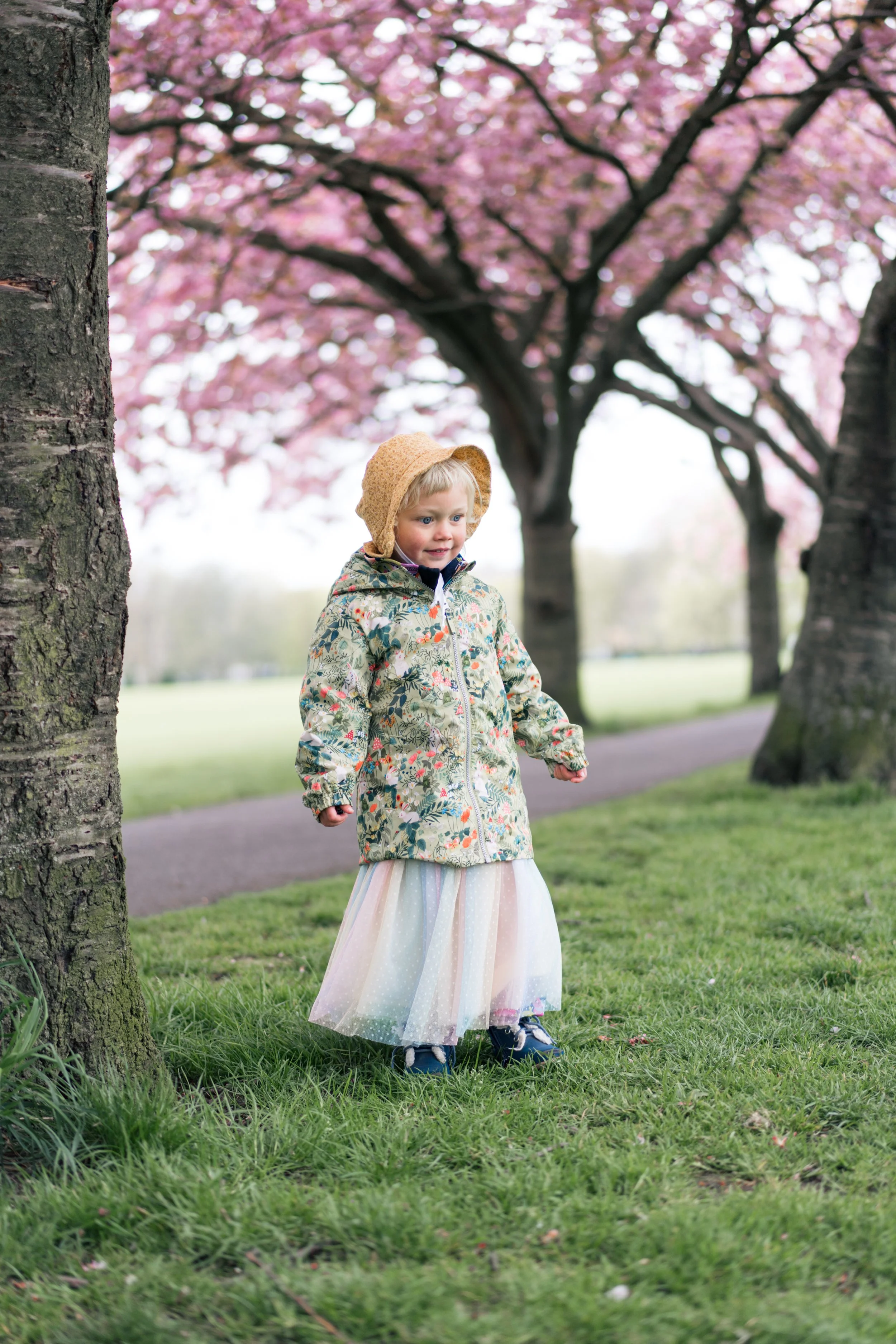 A young girl wearing a floral jacket, tulle skirt, and a yellow hat, standing on grass near trees with pink blossoms, in a park.